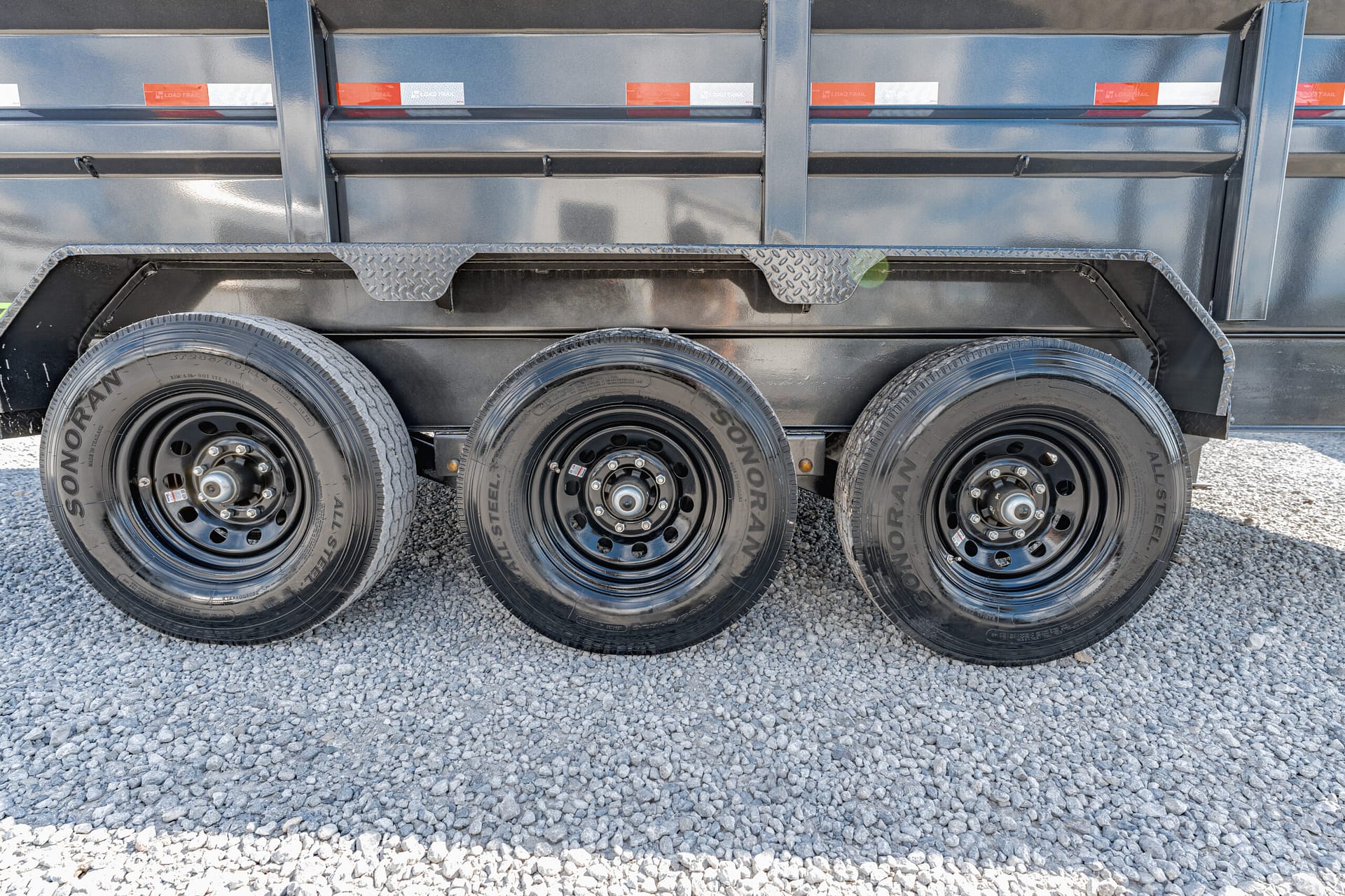 Side view of a heavy-duty trailer showing three axles and black steel wheels on a gravel surface with a metal trailer body above.