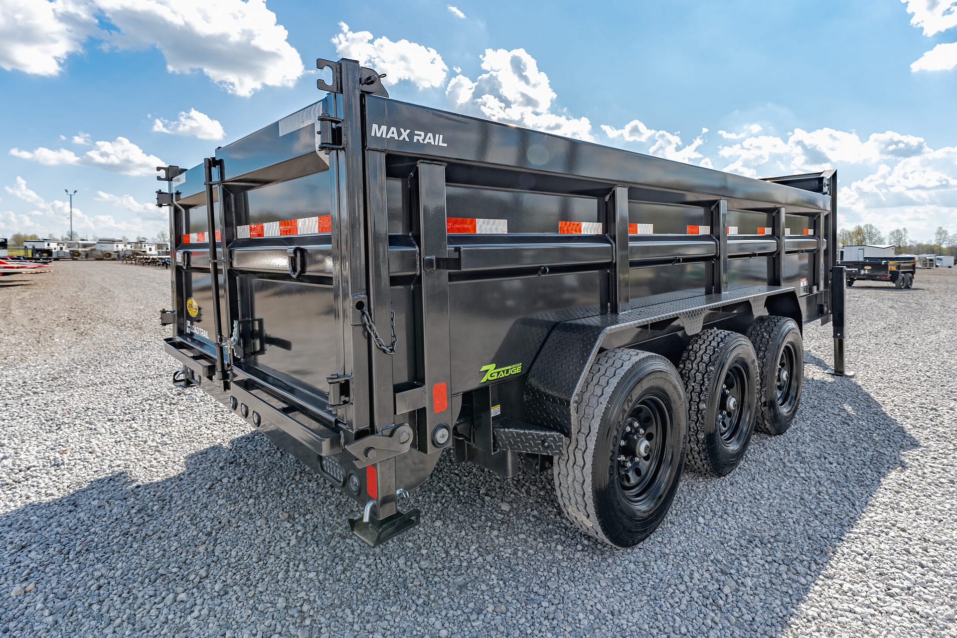 Black Max Rail heavy-duty trailer with tandem wheels on a gravel lot under a blue sky with clouds.