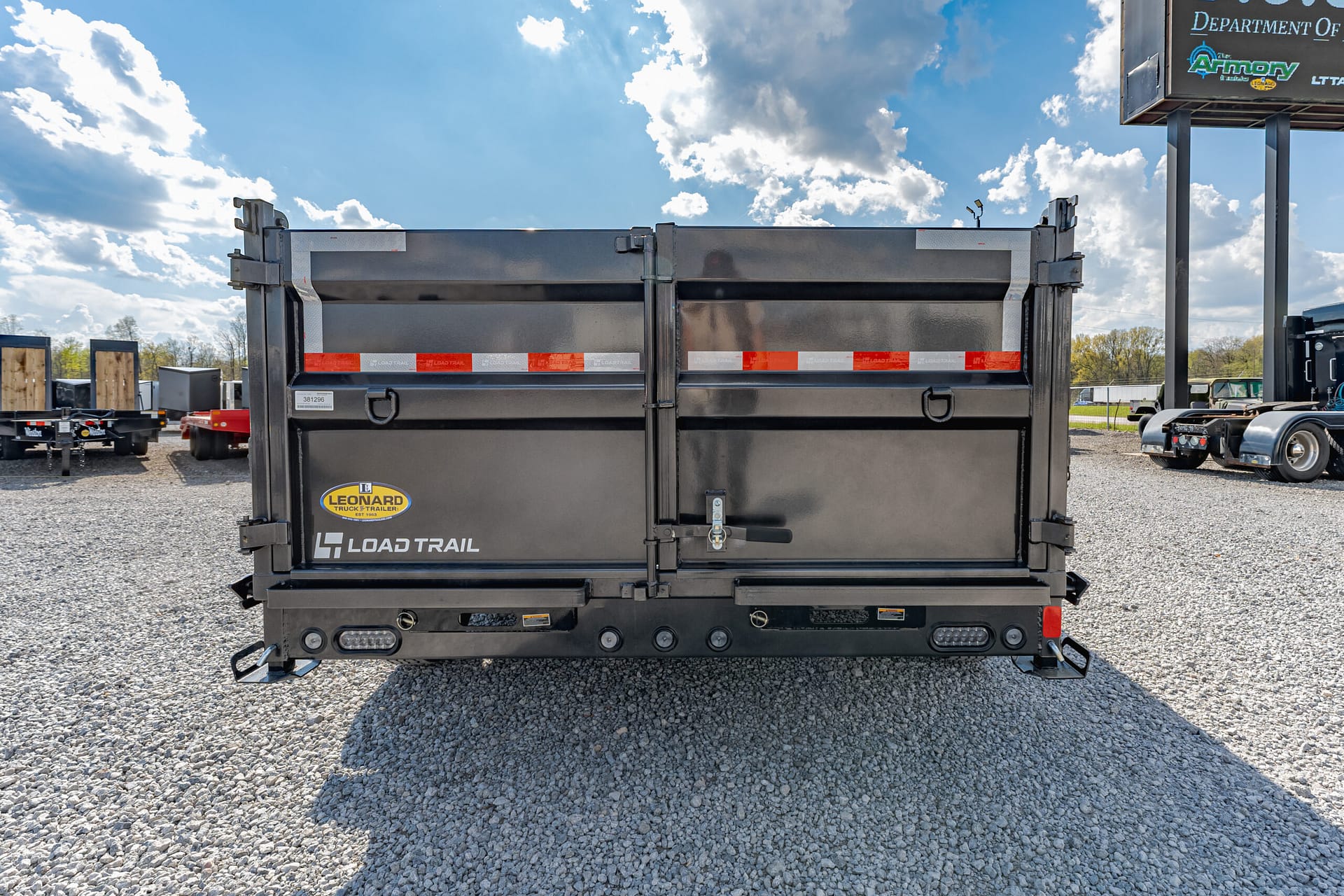 Rear view of a black Load Trail dump trailer with red-and-white reflective tape in a gravel lot, blue sky above with clouds.