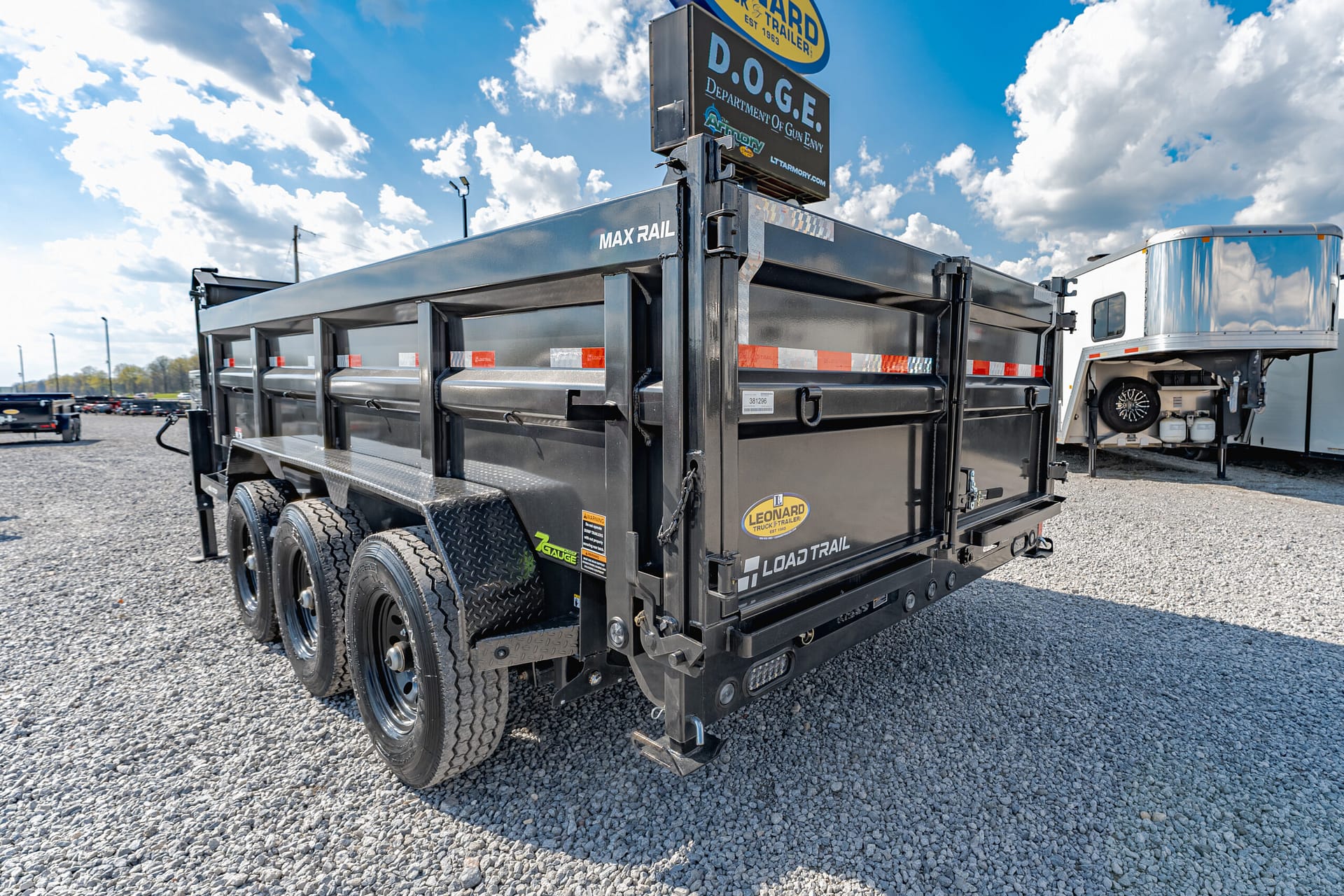 Black heavy-duty dump trailer with multiple axles and orange safety stripes parked on a gravel lot under a blue sky with clouds.