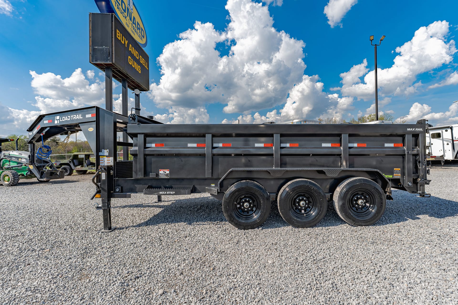 Black Load Trail dump trailer with multiple axles parked on a gravel lot under a bright blue sky with white clouds.