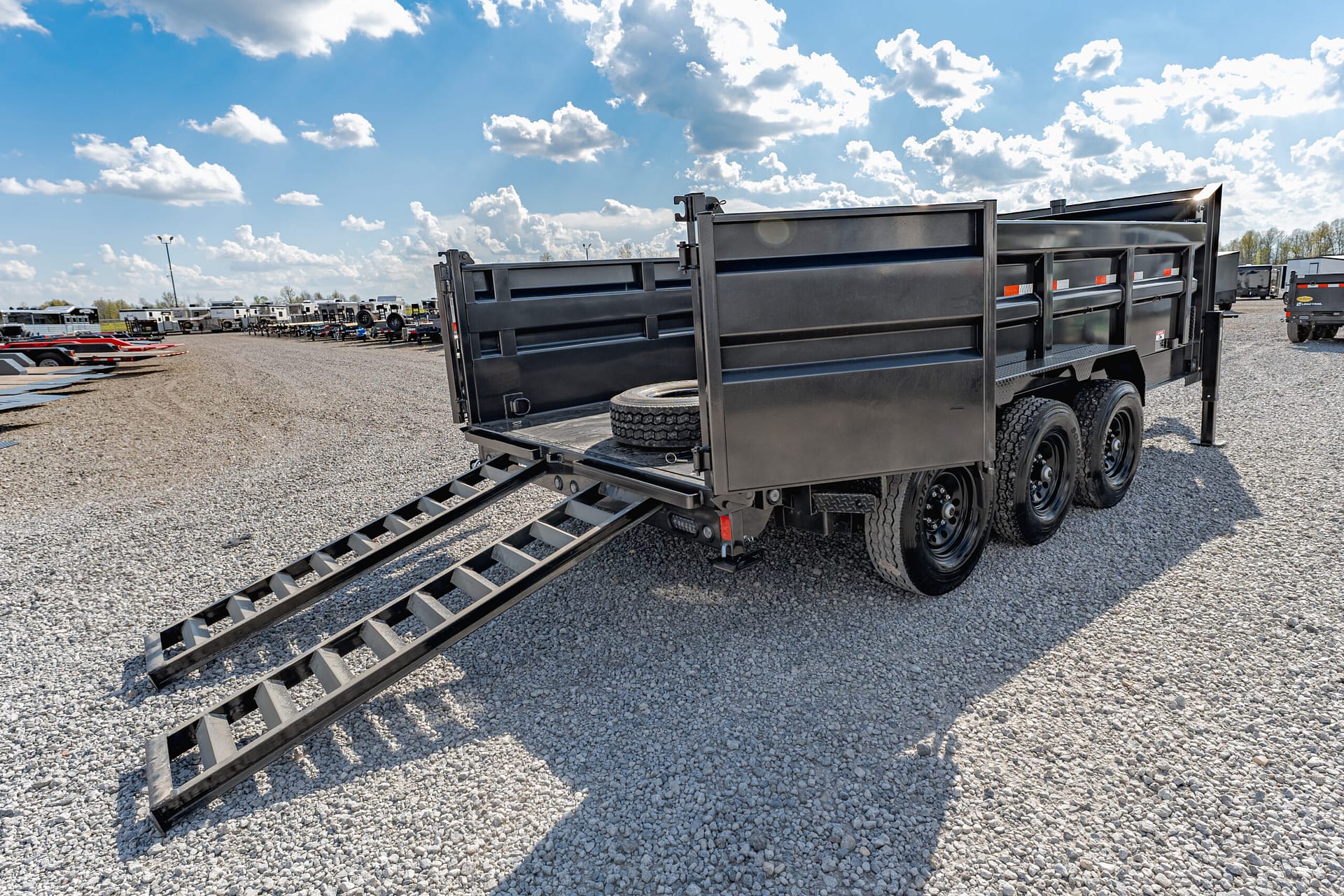 Black dump trailer with fold-down ramps on a gravel lot, spare tire visible on bed and other trailers in the background.