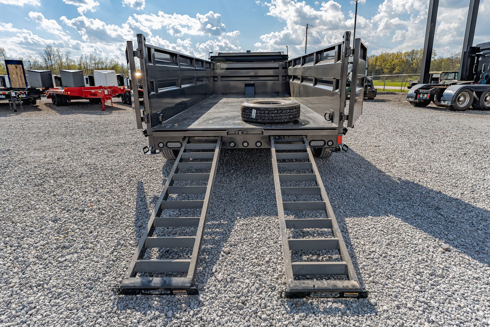 Rear view of a black dump trailer with ramps lowered, a spare tire in the bed, parked on a gravel lot with other trailers in the background