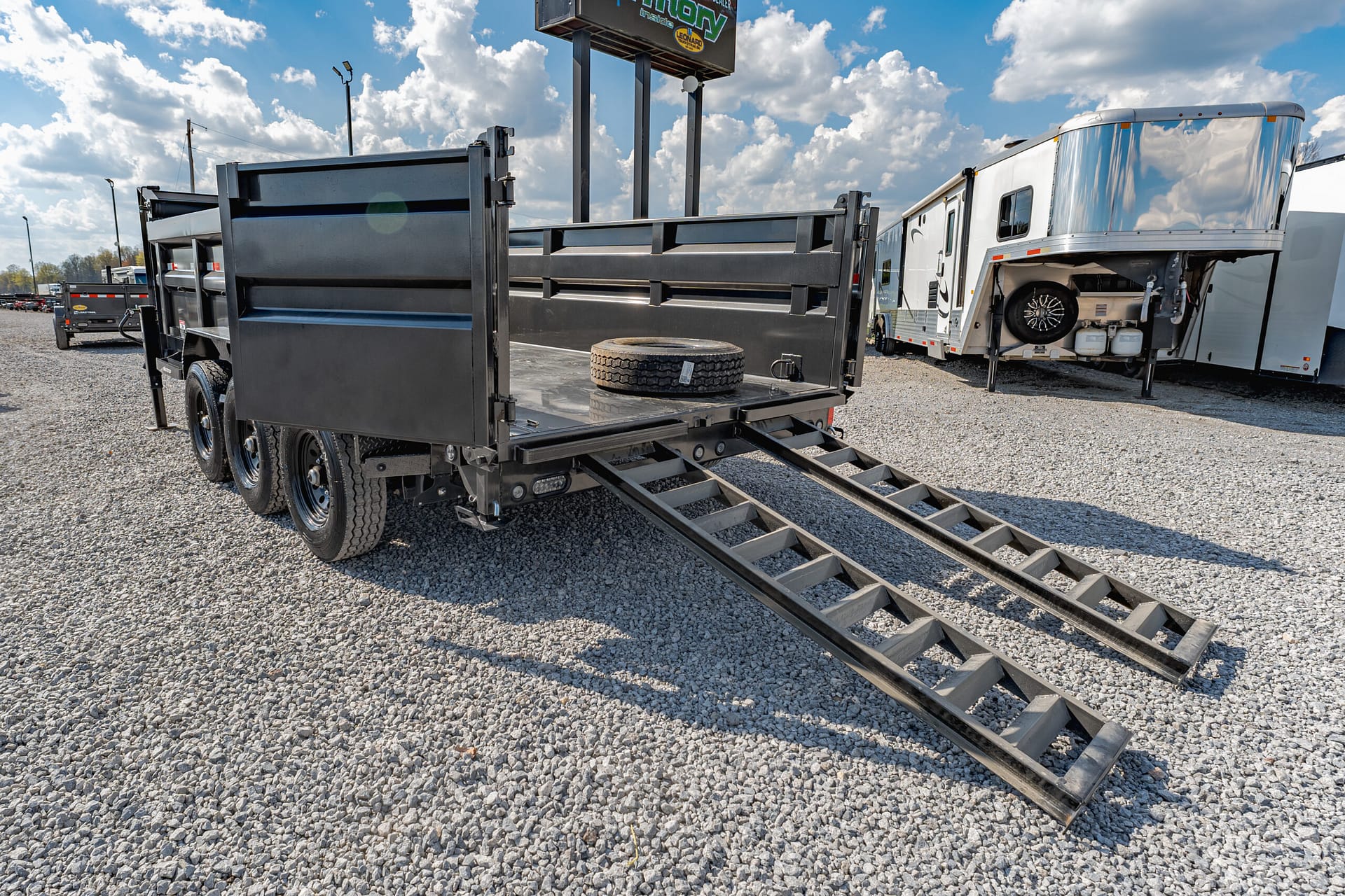 Black steel dump trailer on a gravel lot with ramps extended and a spare tire on the bed, RV trailers in the background.