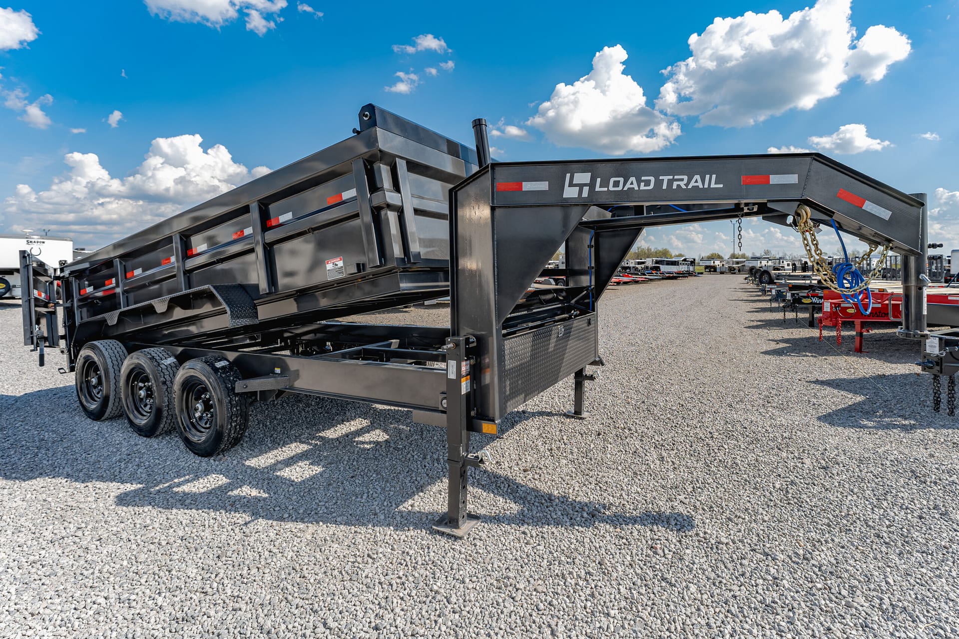 Black heavy-duty trailer with multiple axles and a ramp, parked on a gravel lot beneath a bright blue sky with white clouds (Load Trail logo visible).