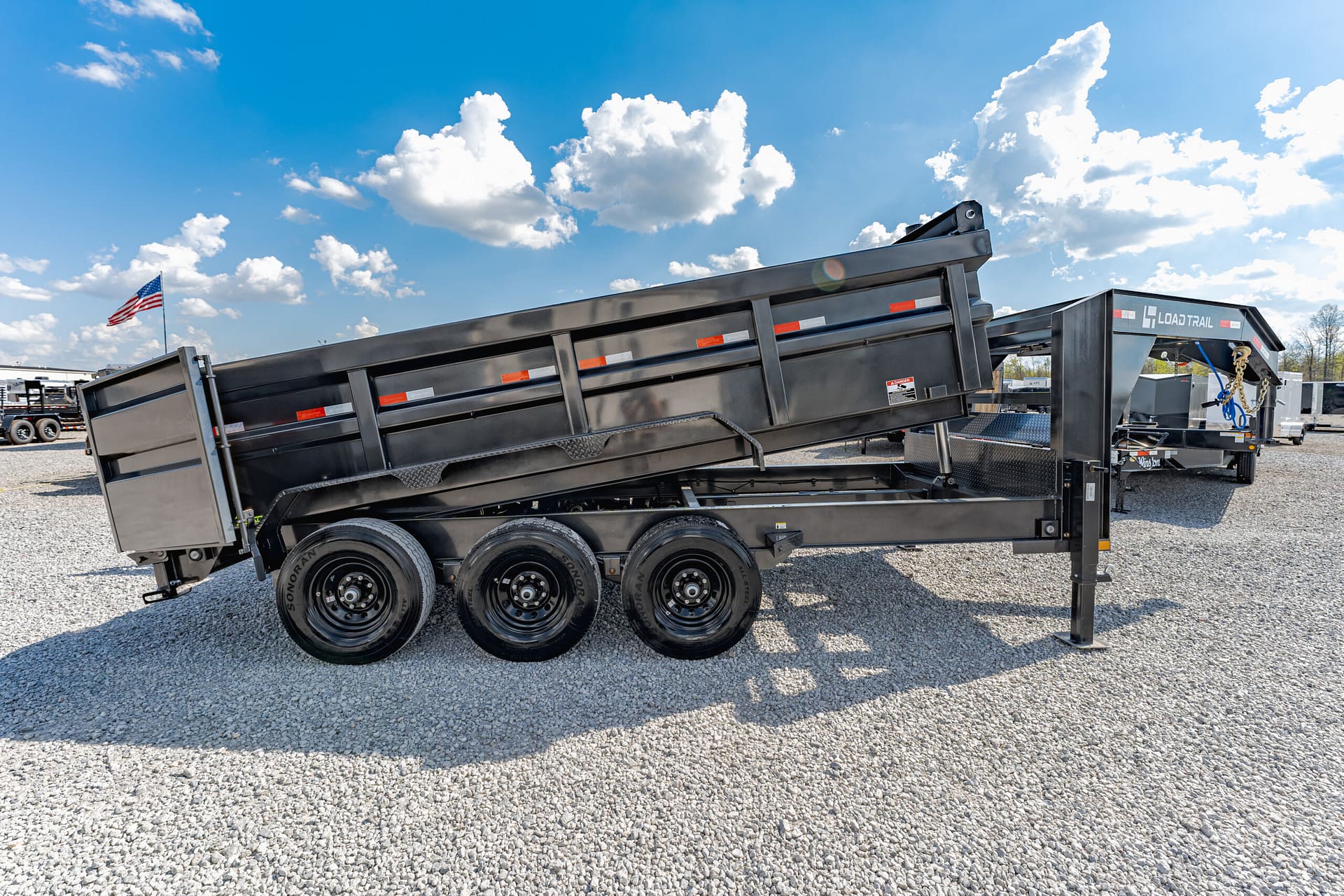 Black dump trailer with raised bed on a gravel lot under a blue, cloudy sky; American flag in the background. (Informative)