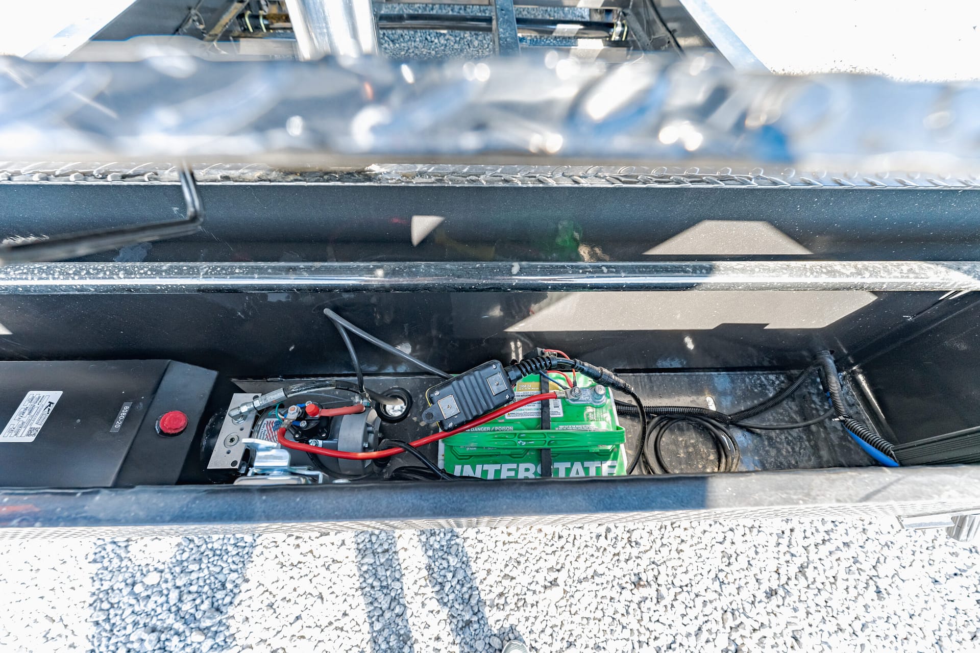 Open vehicle storage area with a green Interstate battery and exposed wiring and connectors inside a metal chassis area, outdoor gravel surface nearby.