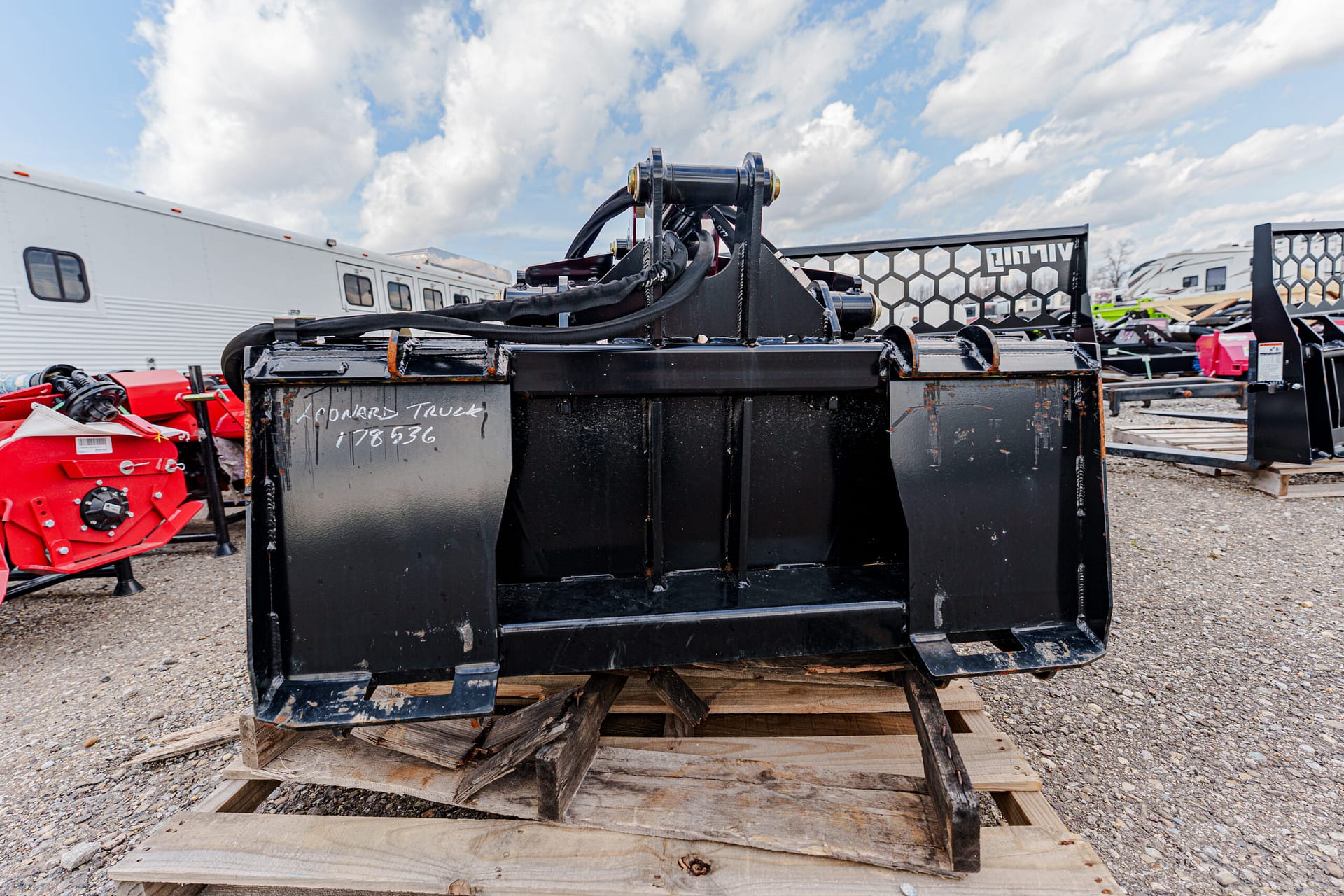 Black metal skid-steer bucket attachment resting on a wooden pallet in an outdoor equipment yard, with hoses and other machinery in the background.