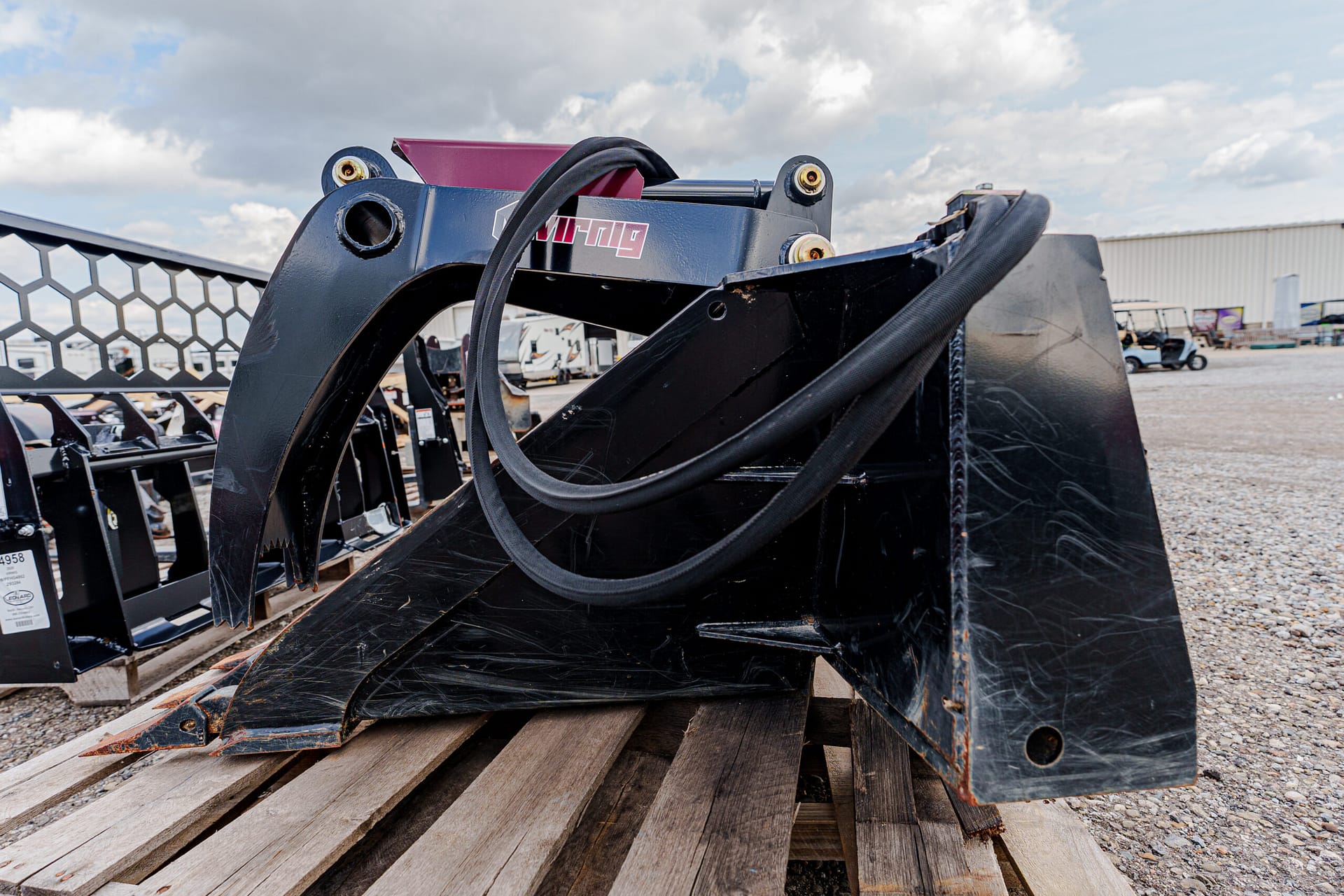 Heavy black skid-steer bucket attachment with hydraulic hoses resting on wooden pallets in an outdoor industrial yard.