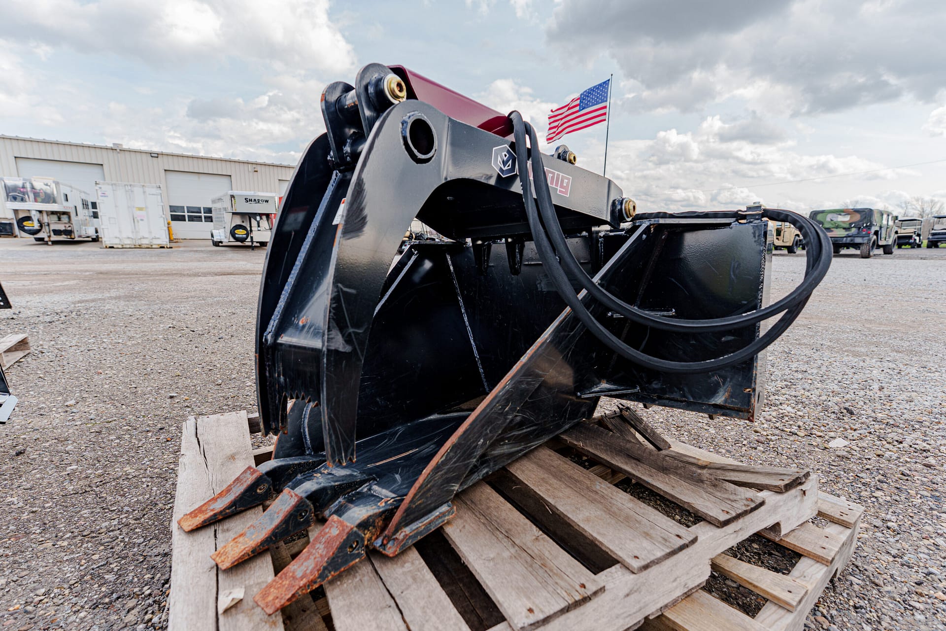 Industrial excavator bucket attachment on a wooden pallet in a gravel yard with a US flag in the background
