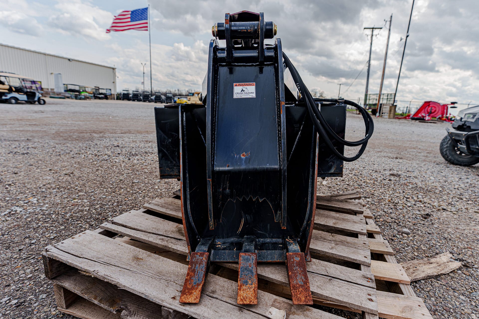 Black excavator bucket with four rusty teeth resting on a wooden pallet in a gravel lot.