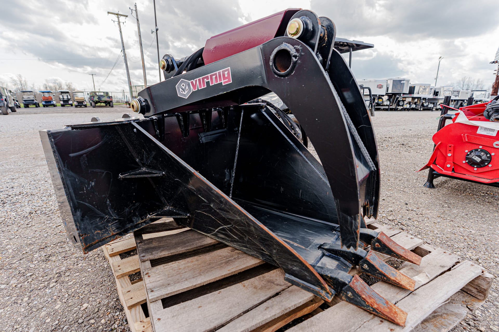 Black heavy-duty loader bucket attachment on wooden pallets in a gravel lot, with other equipment in the background at an outdoor yard.