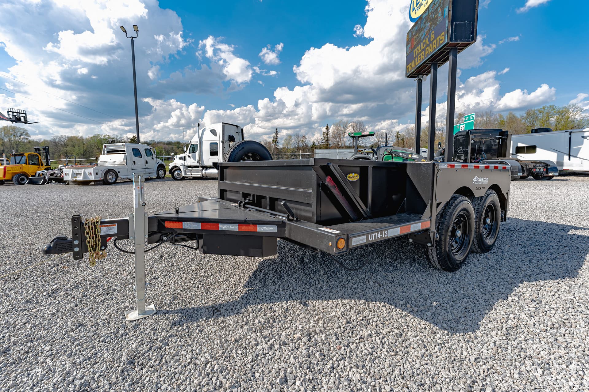 Flatbed trailer in the foreground of a truck yard on a gravel lot, with white semi trucks and a tall sign against a blue sky with clouds in the background.