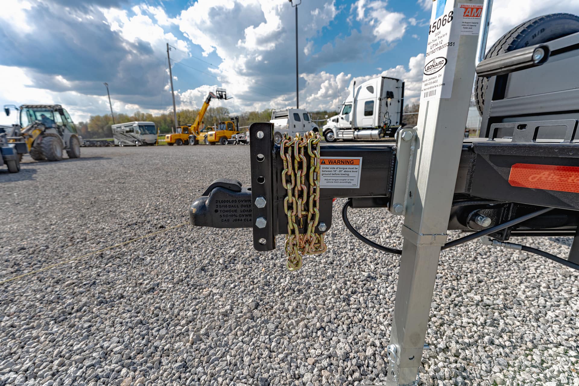 Close-up of a trailer hitch with chains and metal frame in a gravel lot, with trucks in the background under a cloudy sky