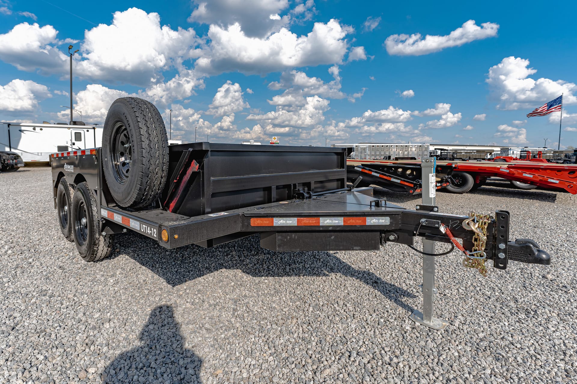Flatbed trailer with a spare tire mounted on the side, parked on a gravel lot under a blue sky with white clouds; an American flag fluttering in the background.