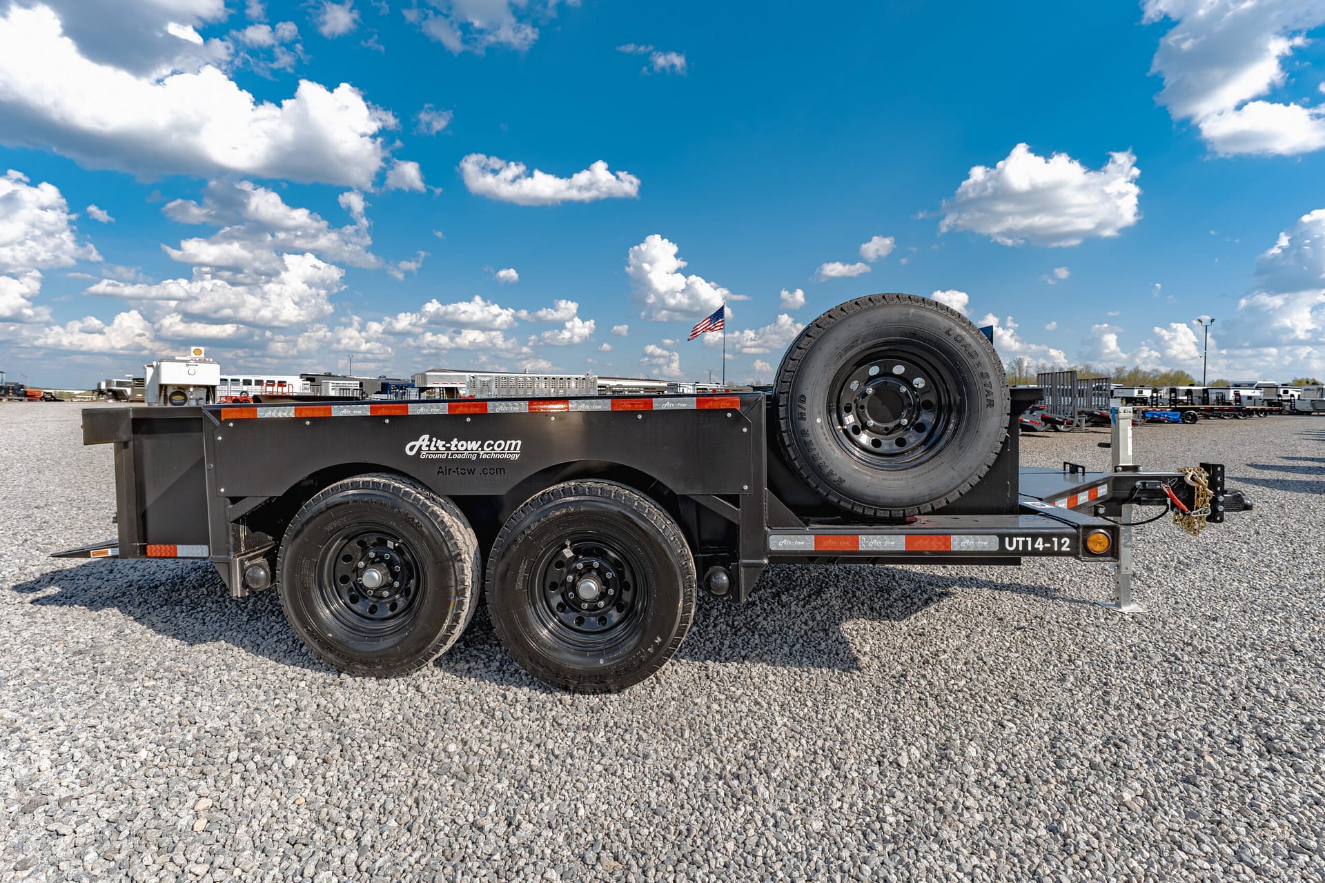 Flatbed trailer with a mounted spare tire in a gravel lot under a blue sky with clouds and distant equipment (functional and informative).