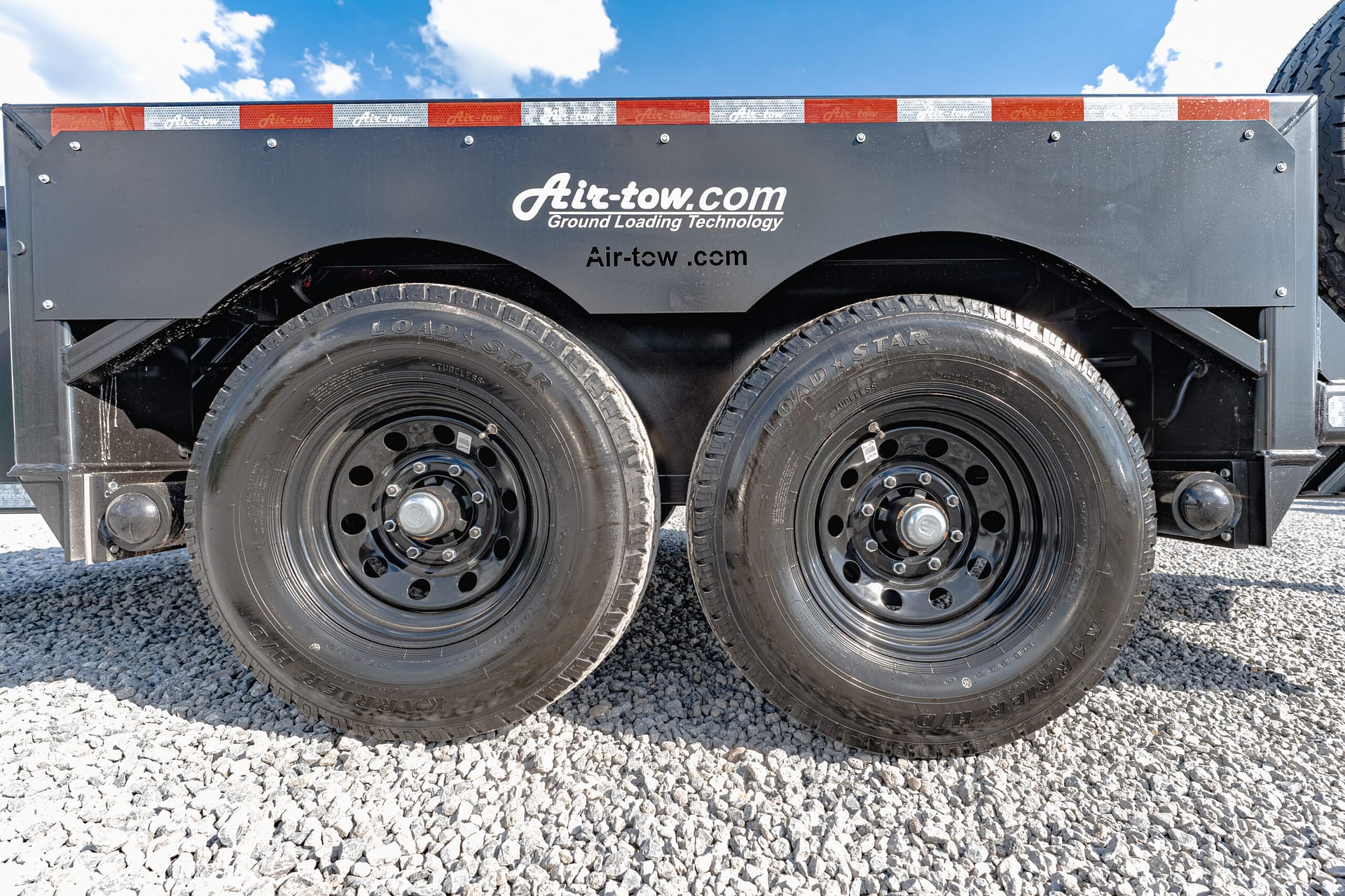 Dual trailer wheels and black fenders on a gray Air-tow trailer with 'Air-tow.com' branding, parked on gravel under a blue sky.