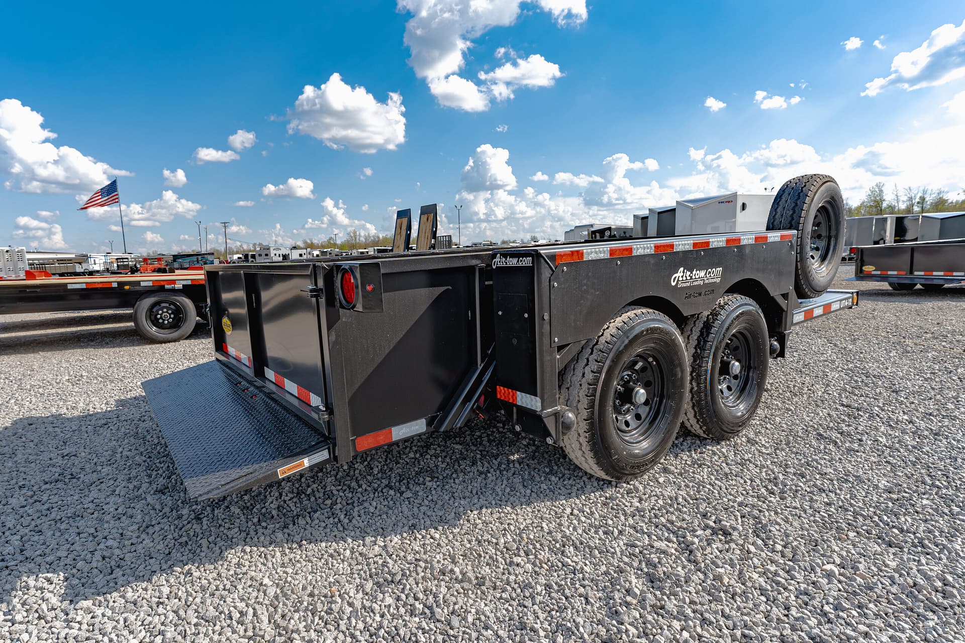 Black flatbed trailer with a mounted spare tire, on a gravel lot under a blue sky with clouds; American flag visible in the distance.
