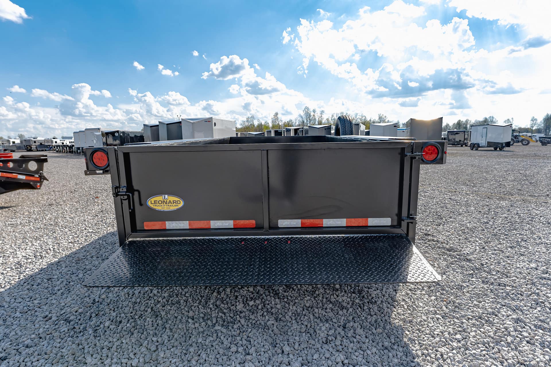 Back of a black utility trailer with red taillights and Leonard Truck & Trailer logo, on a gravel lot with other trailers in the background under a blue sky.