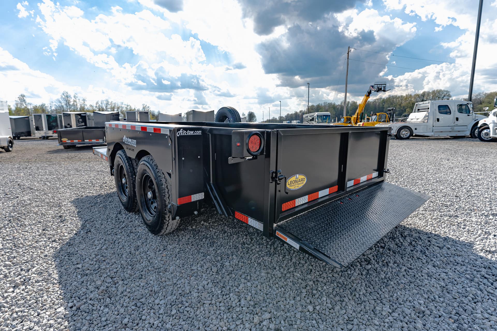 Black flatbed trailer on a gravel lot with other trailers and trucks in the background under a cloudy sky