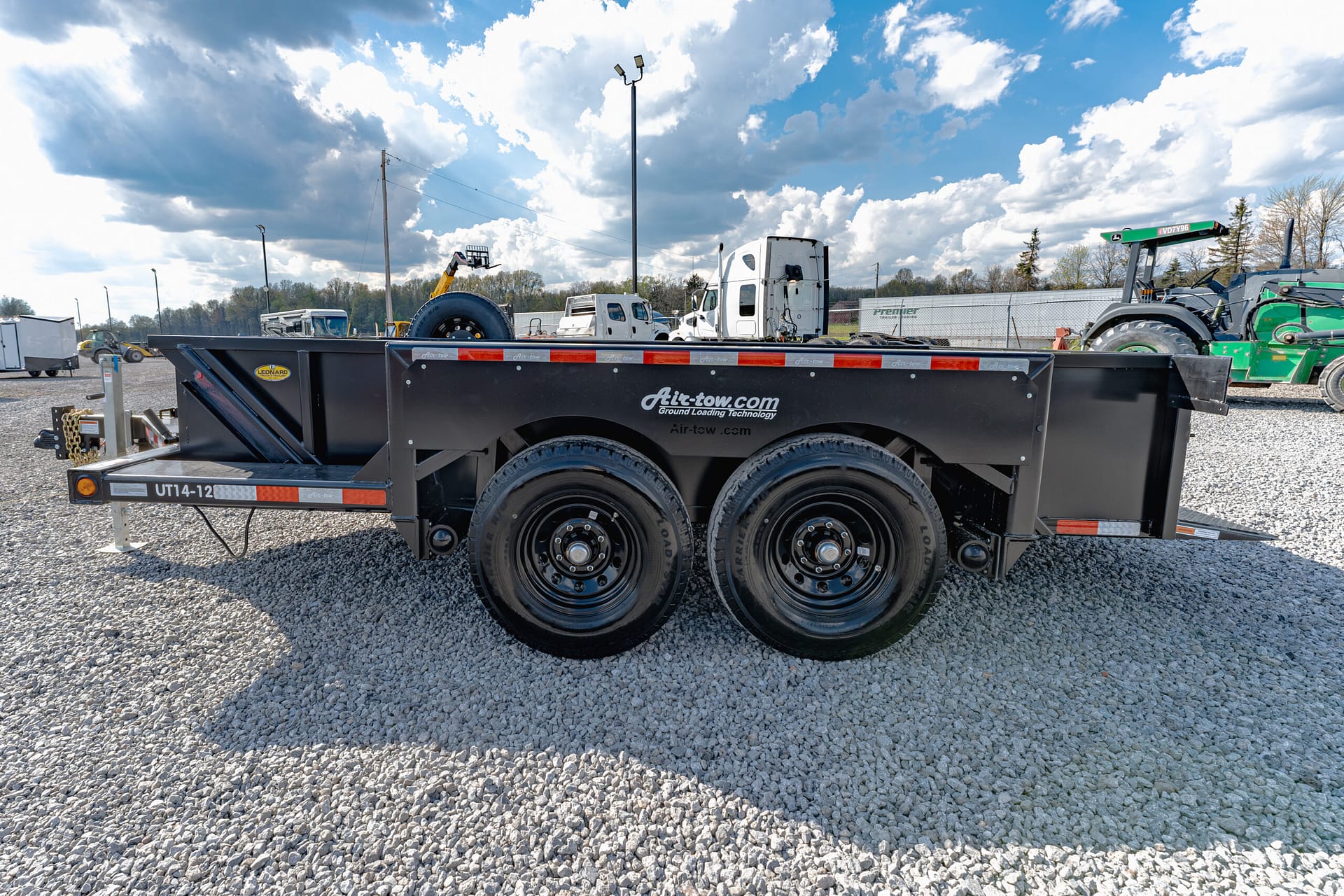 Black flatbed trailer with large dual wheels on a gravel lot, an industrial yard with trucks in the background behind a chain-link fence.