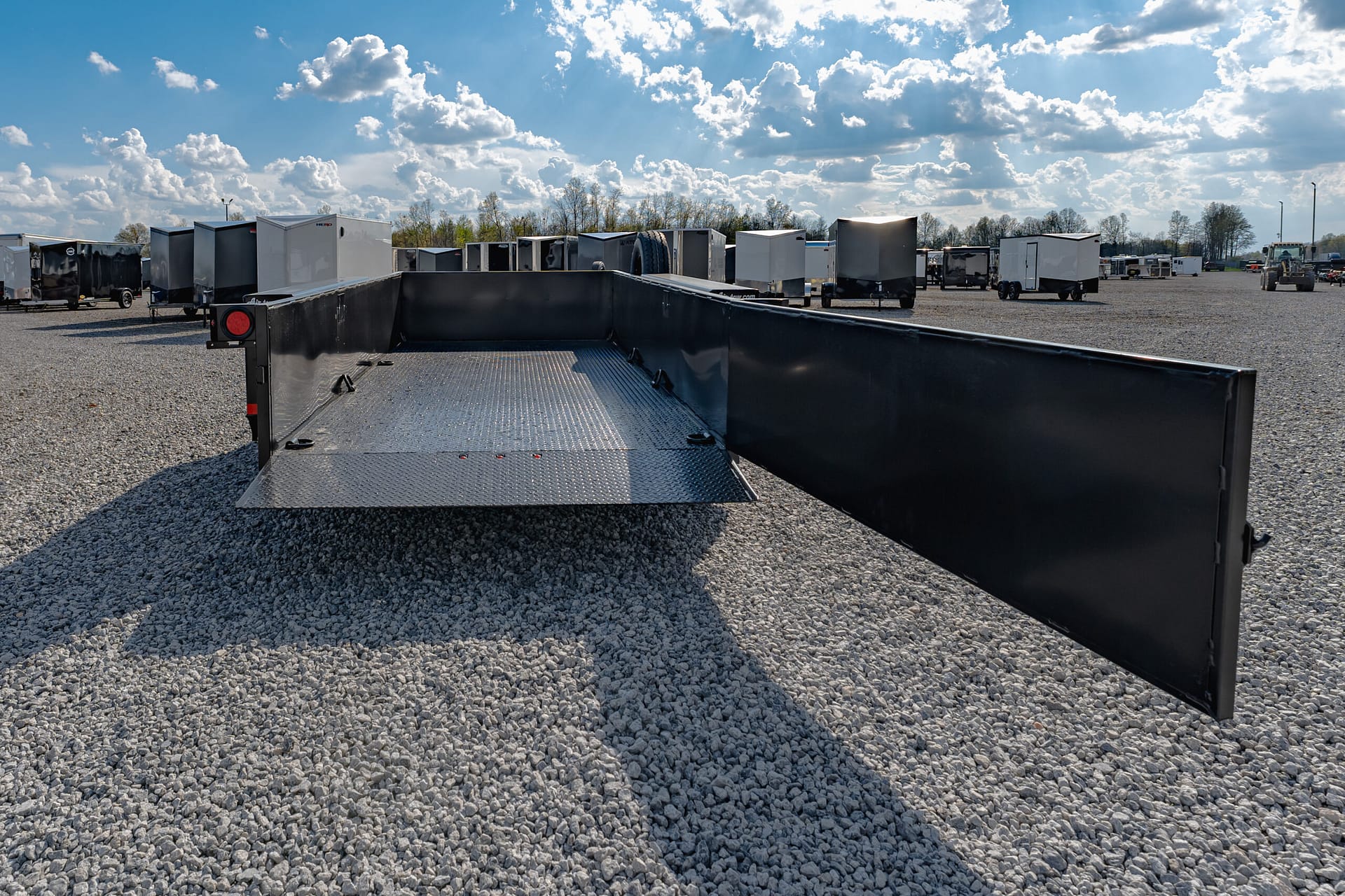 Wide view of a trailer storage lot with numerous enclosed trailers on a gravel surface, seen from the back of a flatbed trailer in the foreground.