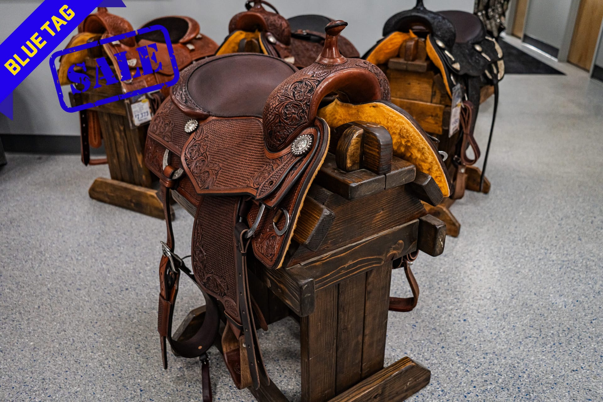 Leather Western saddles with intricate tooling on a wooden rack, a blue 'Blue Tag' sale banner in the corner.
