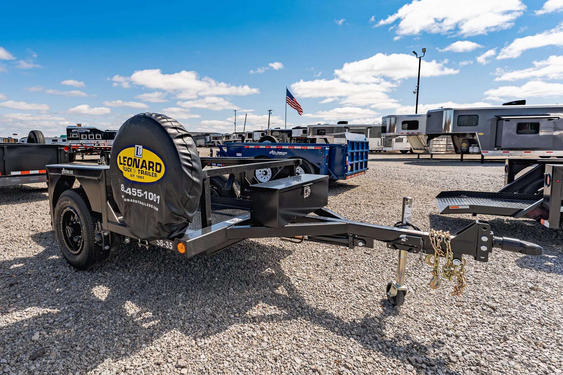Close-up of a black trailer with a spare tire cover reading Leonard Truck & Trailer in the foreground, row of trailers and RVs in the gravel lot under a blue sky.