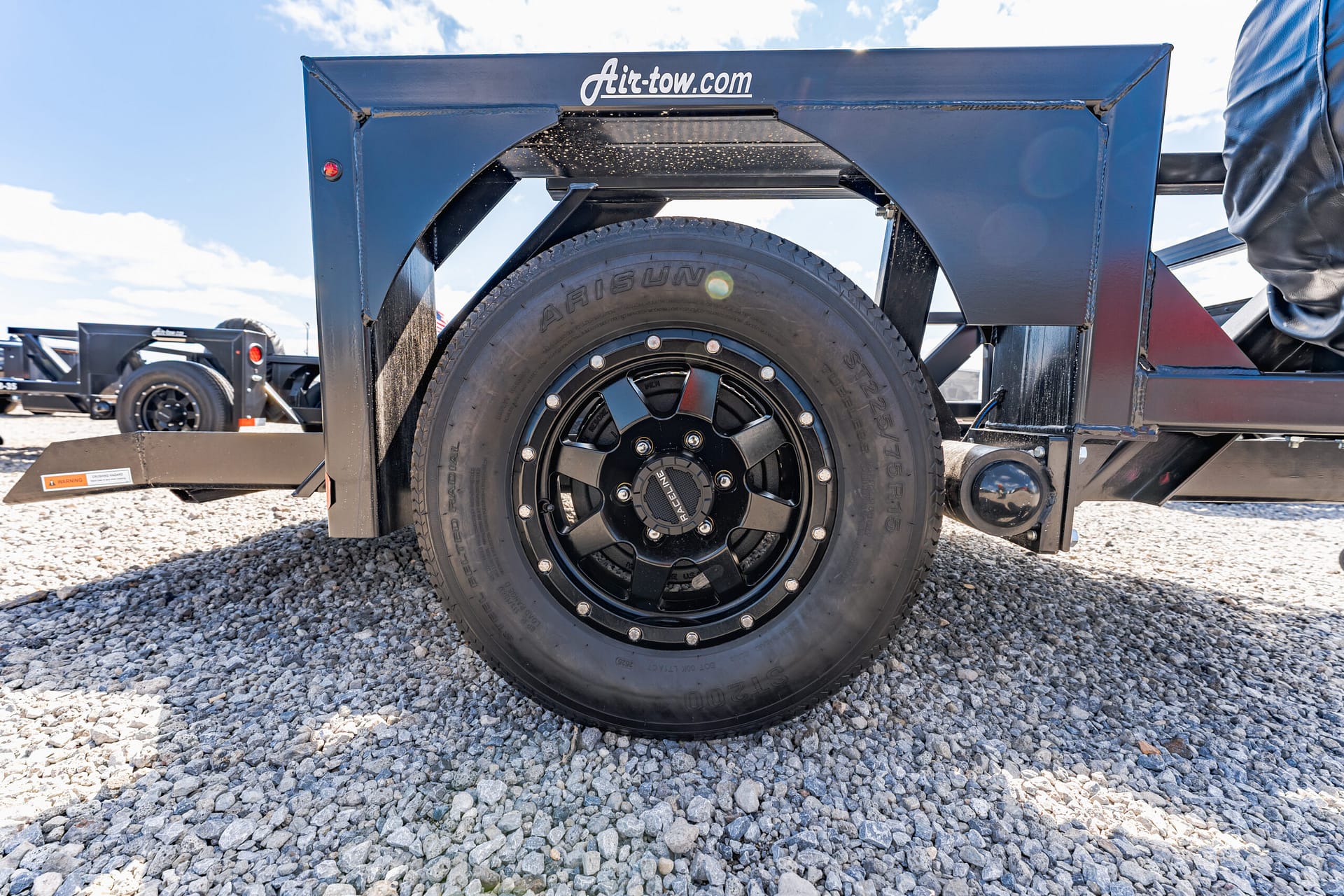 Close-up of a black trailer wheel and tire mounted in a blue metal frame above a gravel surface, with 'Air-tow.com' branding visible on the frame.