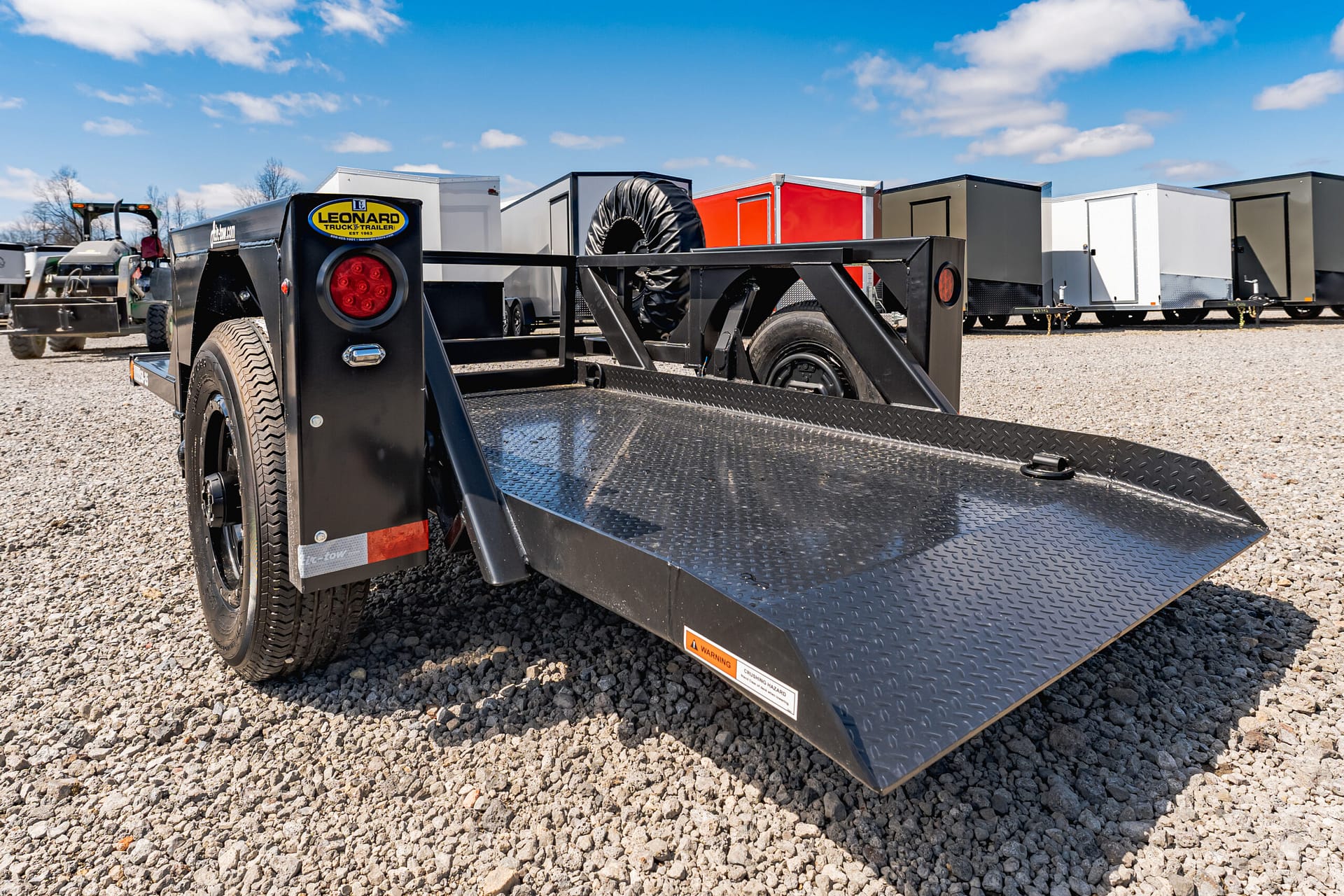 Close-up of a black metal trailer ramp with a diamond-plate surface on a gravel lot, with other trailers in the background under a blue sky.