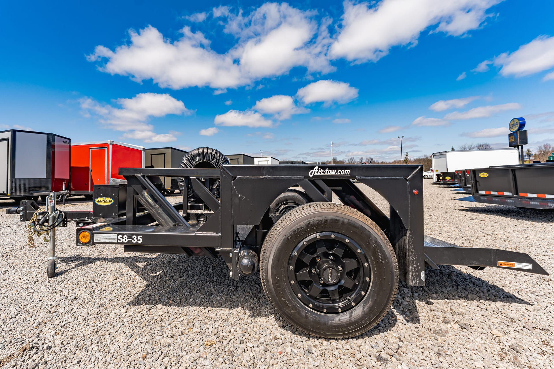 Black Air-tow trailer frame and large wheel on a gravel lot under a bright blue sky with white clouds.