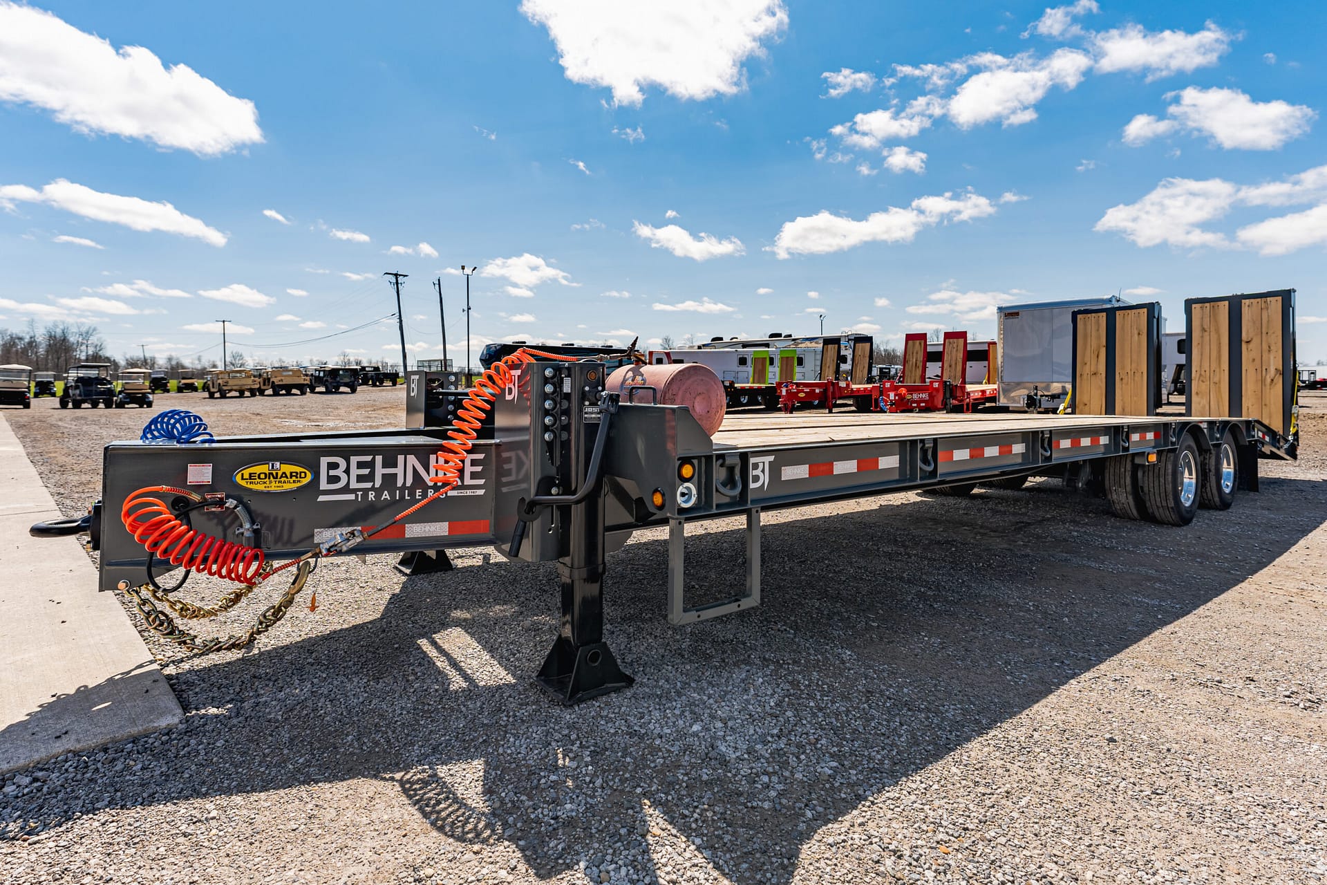 Flatbed trailer with orange coiled hoses, industrial equipment, and wooden ramps in a truck yard under a blue sky.