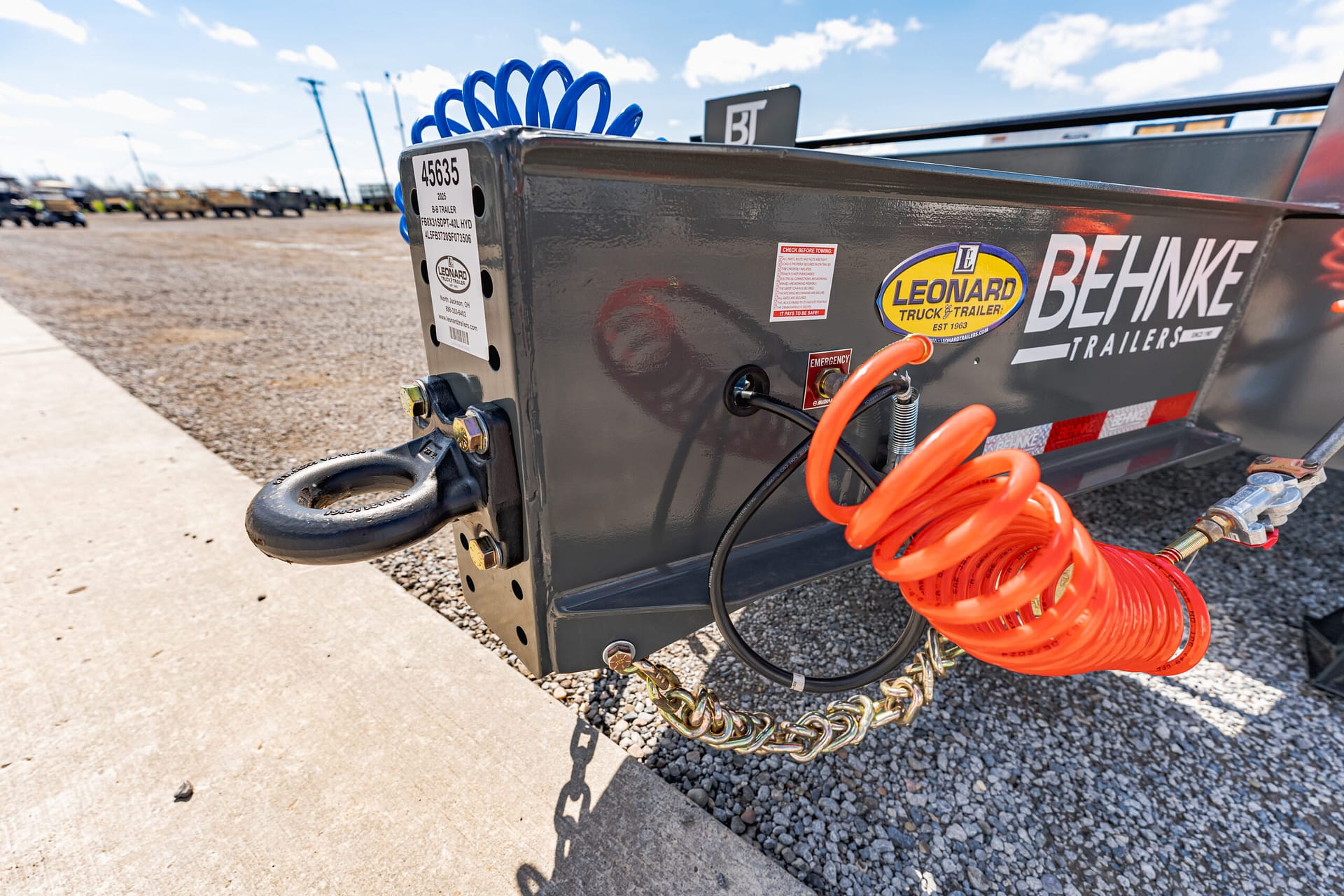 Close-up of a trailer hitch with orange air hoses and a Leonard trailer decal on a gray trailer body, parked on gravel.