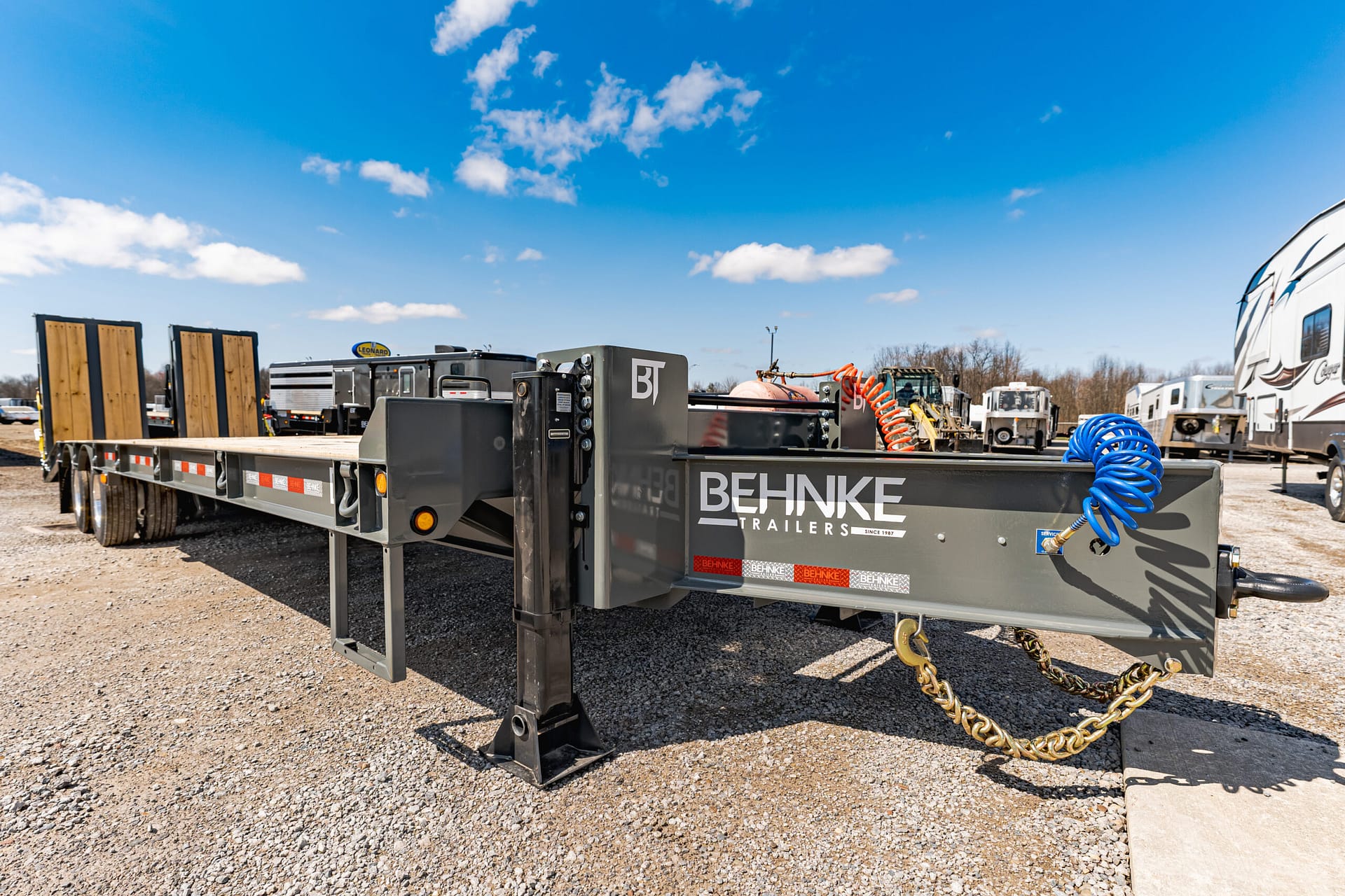 Wide shot of a Behnke Trailers flatbed in a gravel lot with a blue sky and equipment nearby