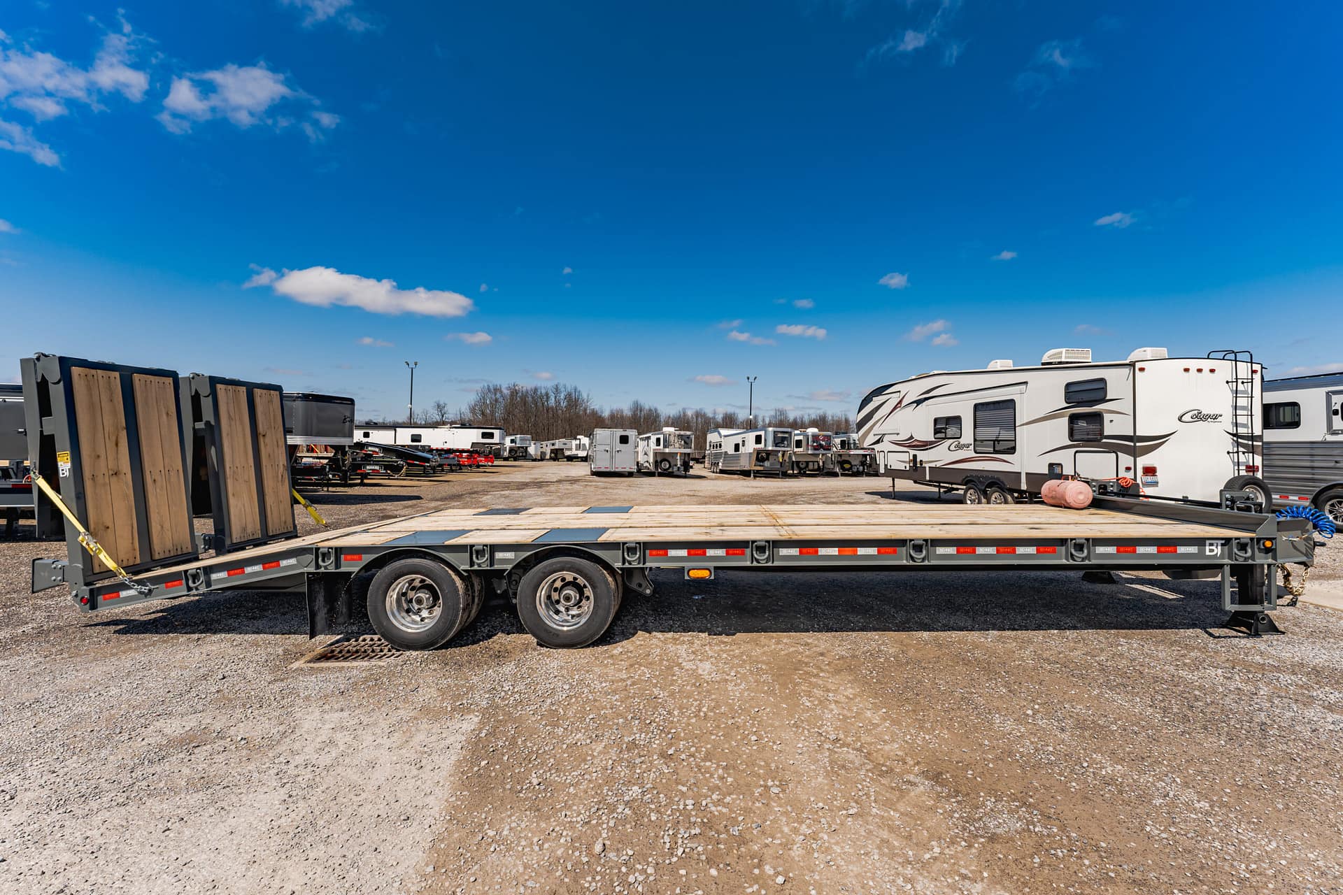 Flatbed trailer in the foreground with stacked wooden boards on the left and a row of RVs parked in a gravel lot under a bright blue sky.