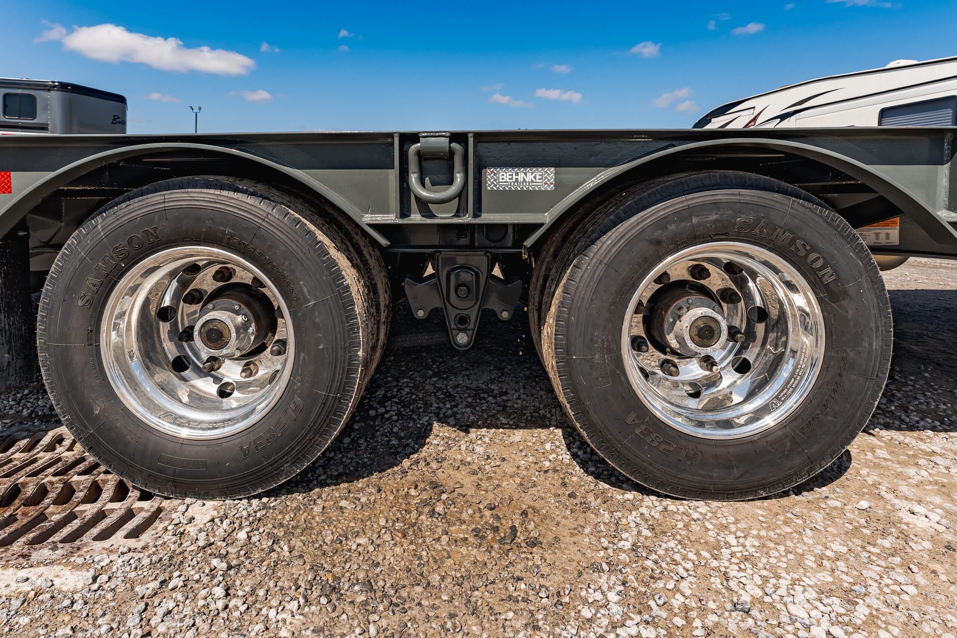 Close-up of dual trailer wheels with chrome rims, showing the trailer’s undercarriage on a gravel surface. A Behnke Trailers plate is visible between the axles, under a blue sky.