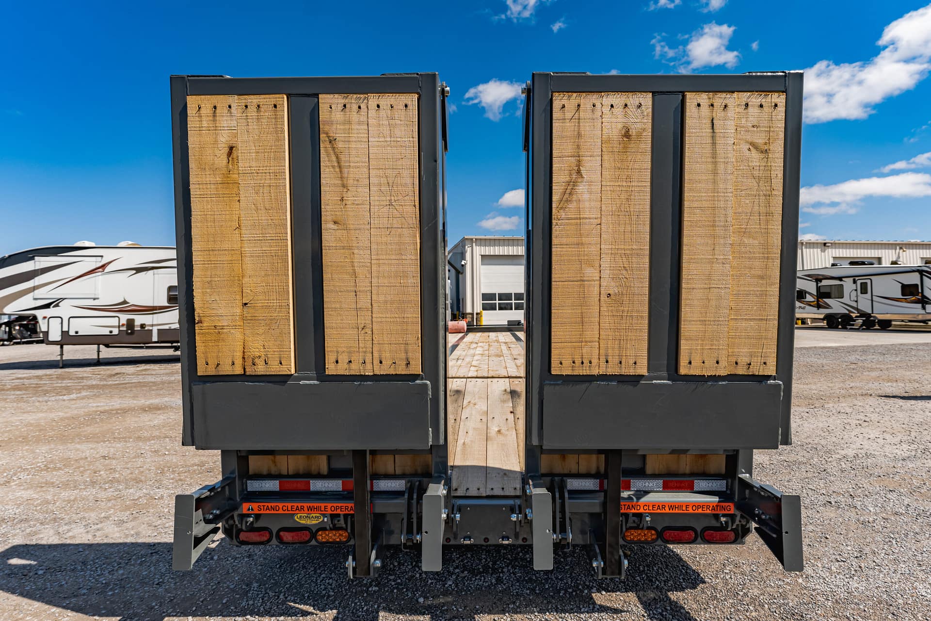 Rear view of two flatbed trailers with weathered wooden side panels and orange safety text, RVs in the background.
