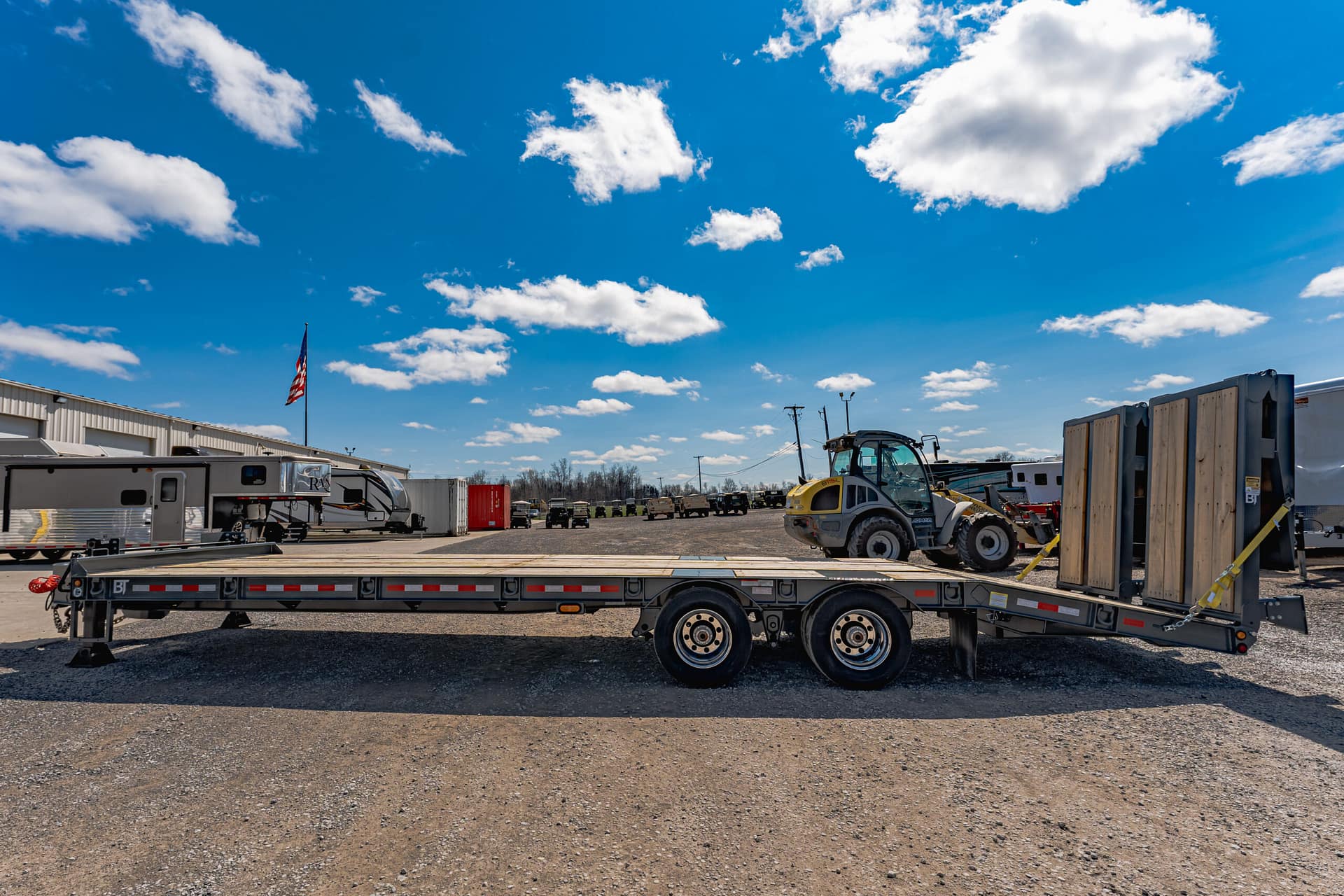 Outdoor industrial yard scene with a long flatbed trailer in the foreground, RVs and equipment nearby, under a bright blue sky with scattered white clouds.