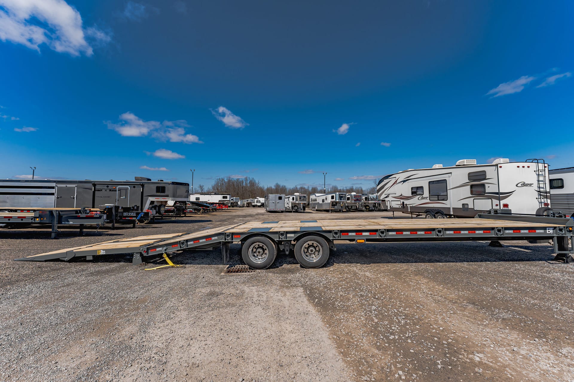 Row of travel trailers and large trailers parked in a gravel lot under a bright blue sky with a few clouds, with a flatbed ramp in the foreground.
