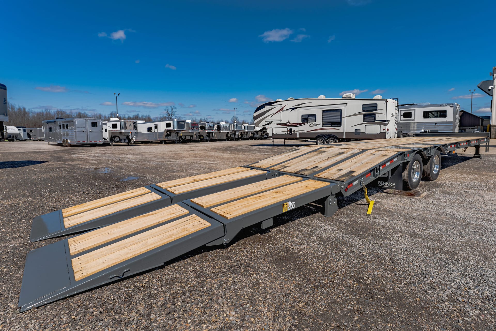Line of white RVs and campers parked in a gravel lot, with a flatbed trailer and wooden ramps in the foreground.