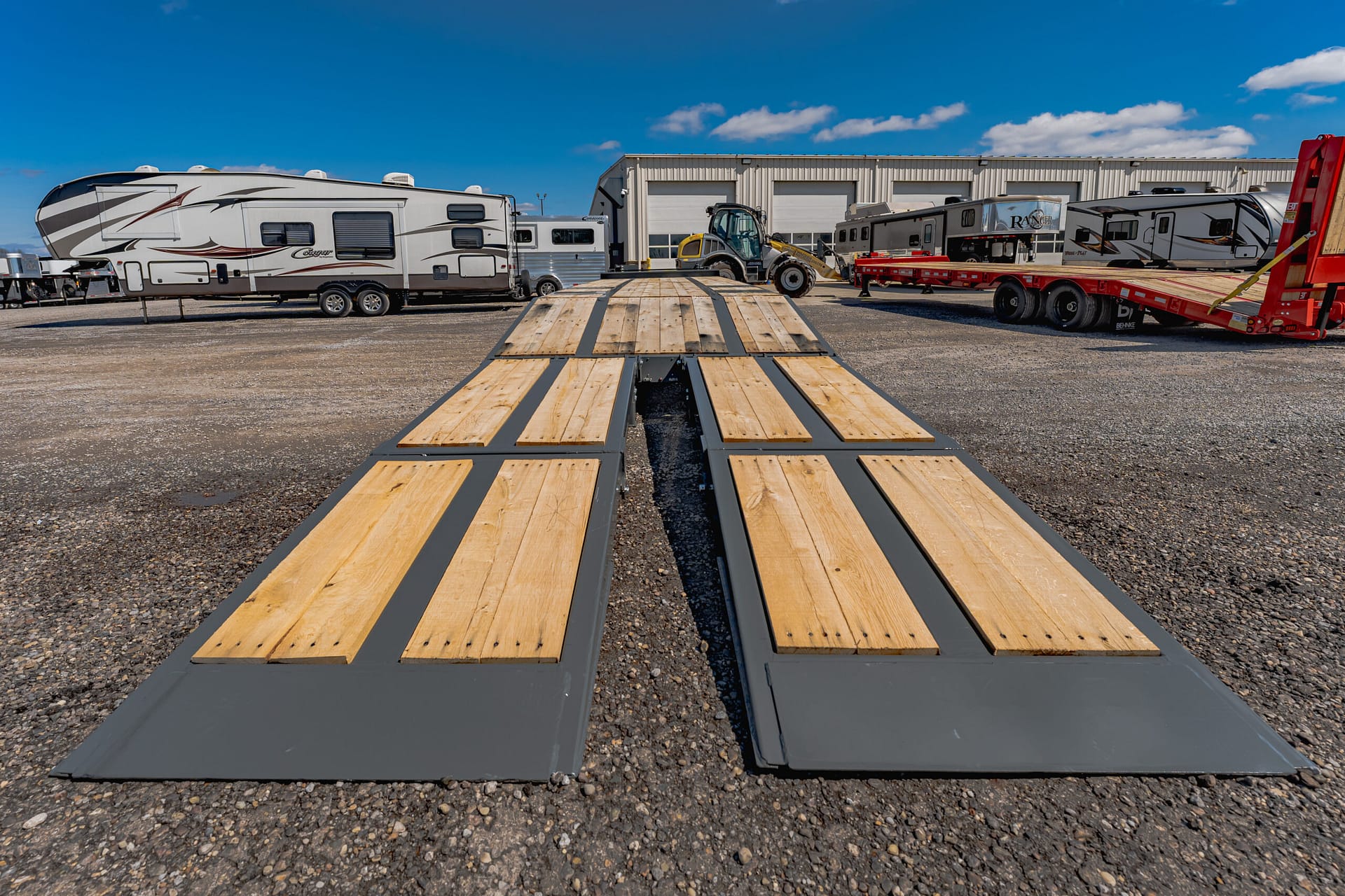 Wide view of a wooden flatbed trailer in the foreground with RVs and a service building under a blue sky.