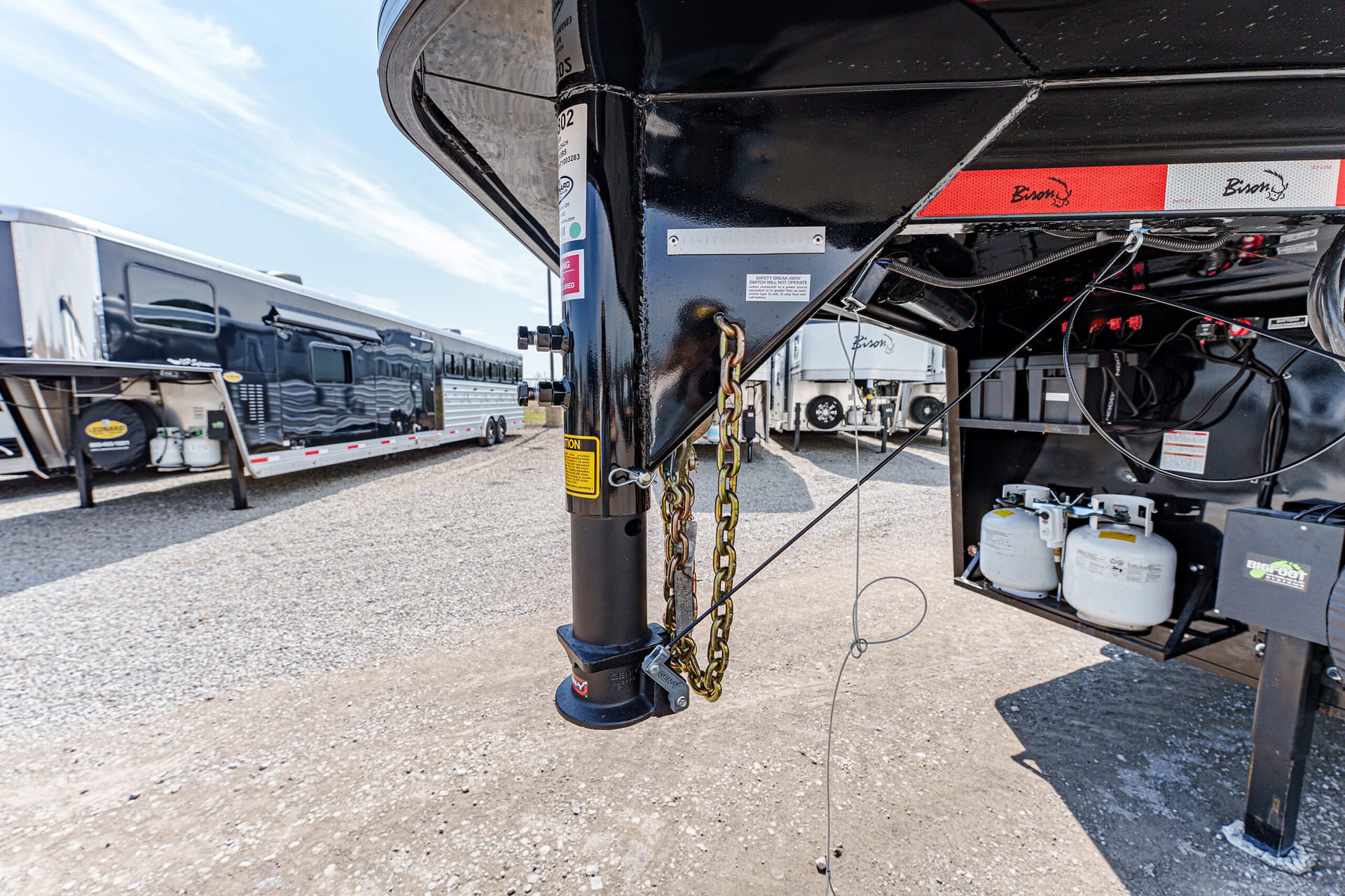 Close-up of the hitch on a black travel trailer, showing a yellow safety chain and a propane-tank storage area nearby in a gravel lot.
