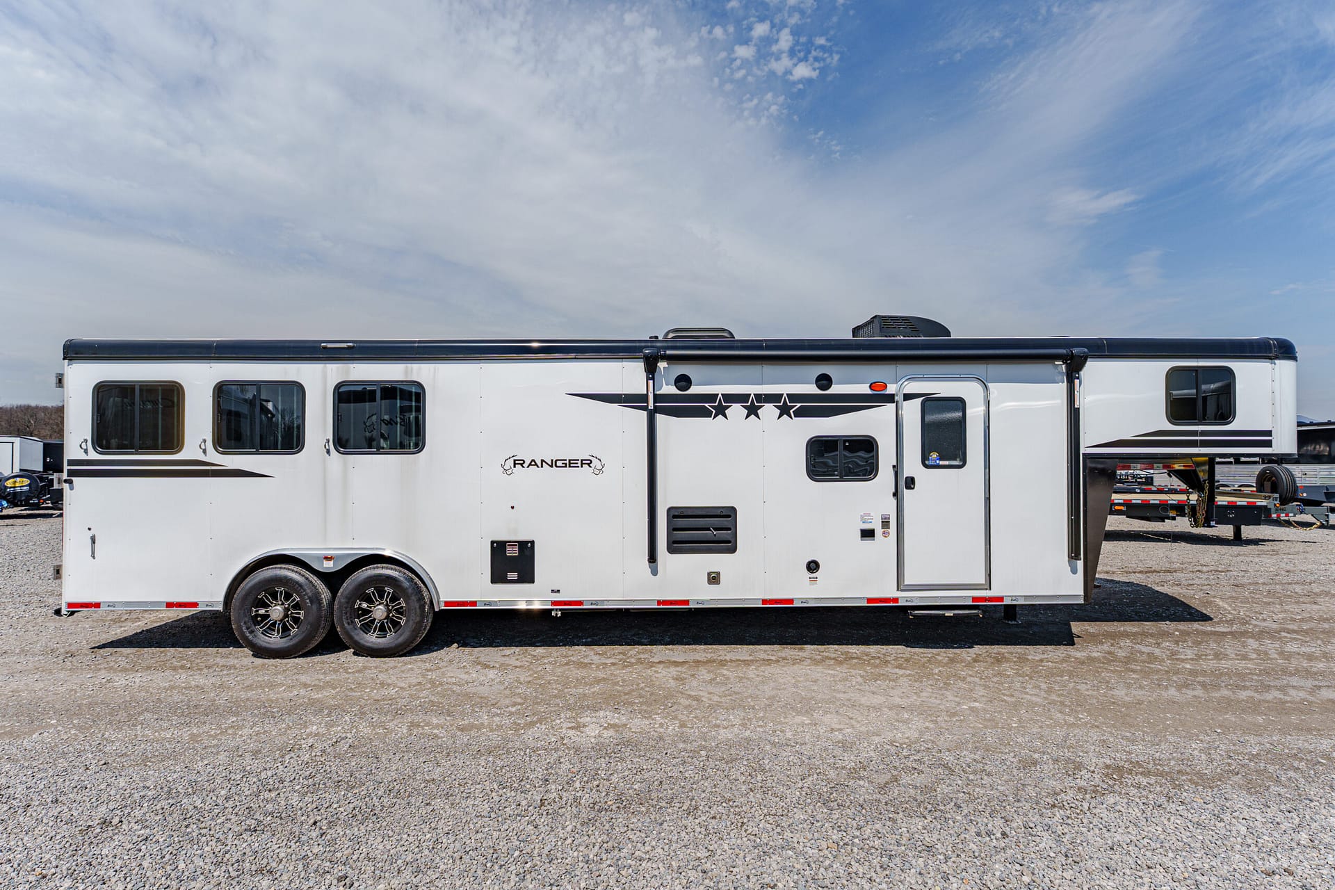 White long RV trailer with black trim and star decals parked on a gravel lot under a blue sky, two-axle chassis with windows along the side.