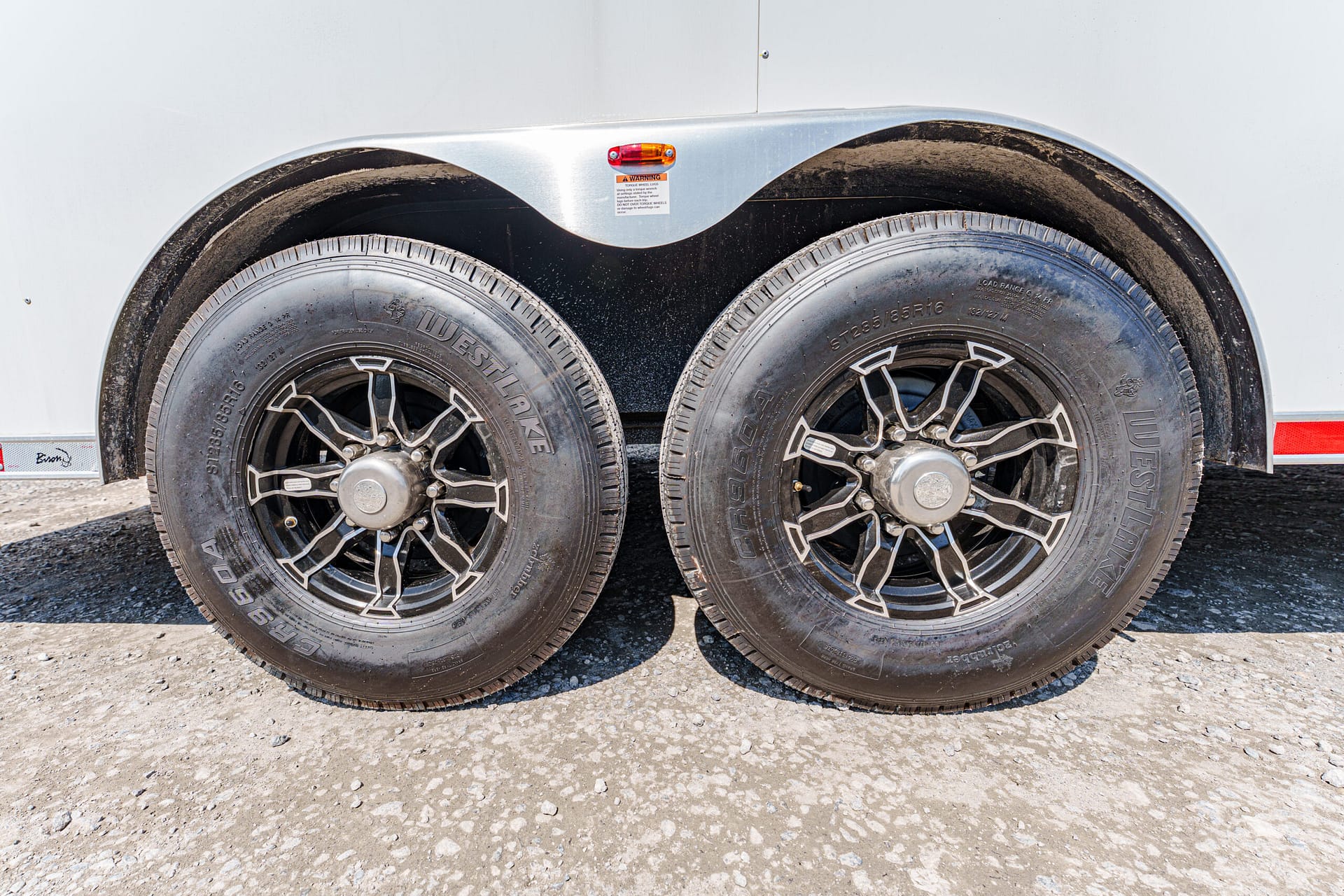 Two trailer wheels side by side with black alloy rims on a gravel surface.