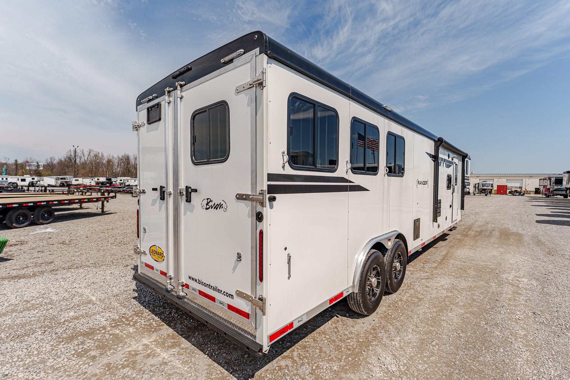 White Bison horse trailer with rear double doors and side windows, parked on a gravel lot with other trailers nearby.
