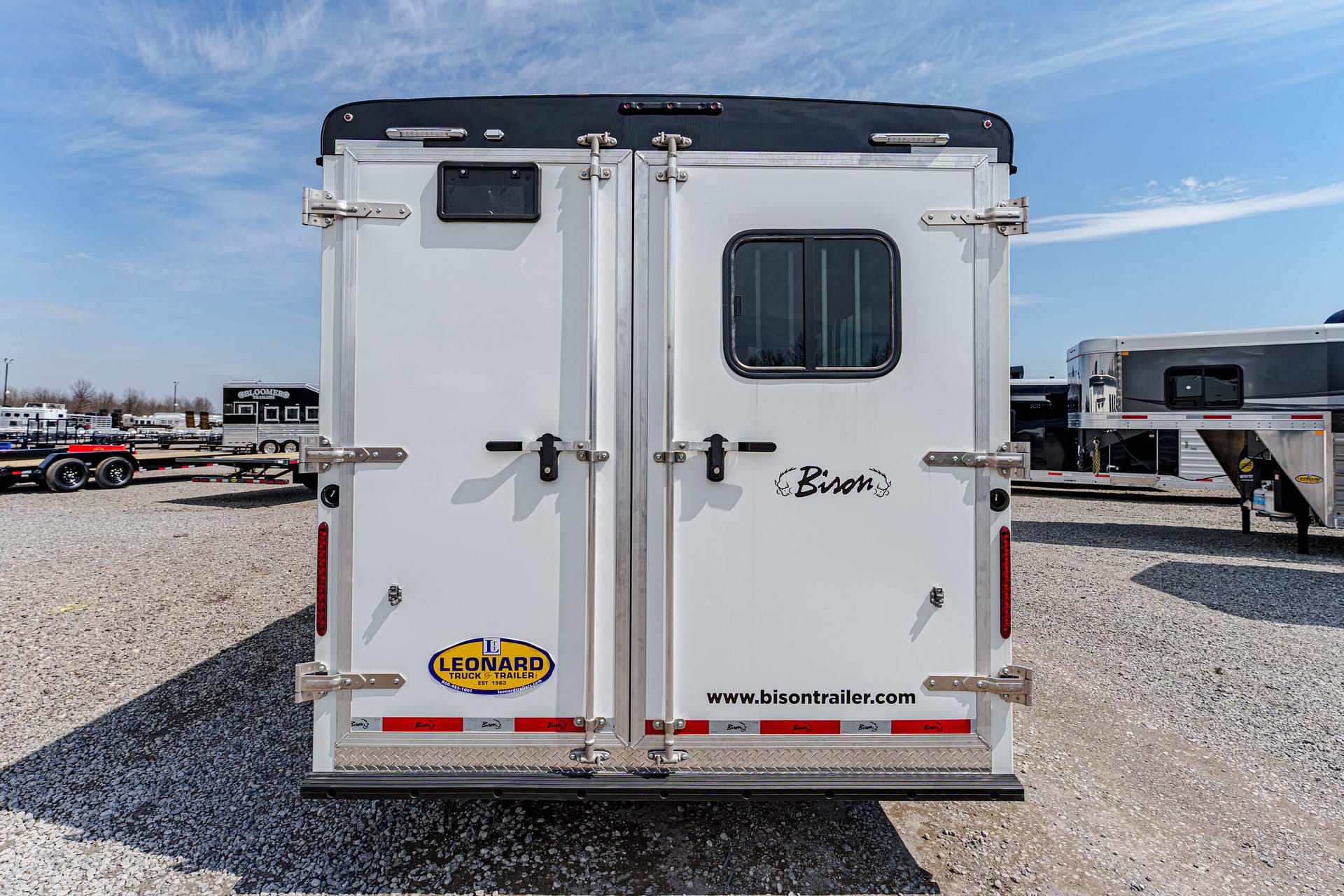 Back of a white Bison horse trailer with twin doors, small window, and branding stickers visible against a gravel lot.