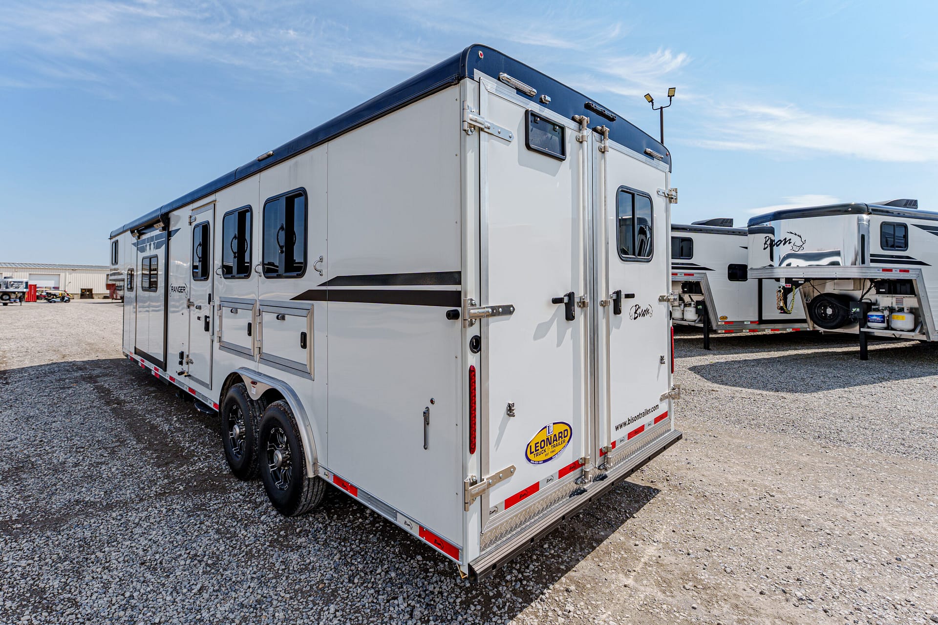 White gooseneck horse trailer with two axles and blue trim parked on a gravel lot, with other trailers in the background; features multiple windows and rear double doors.