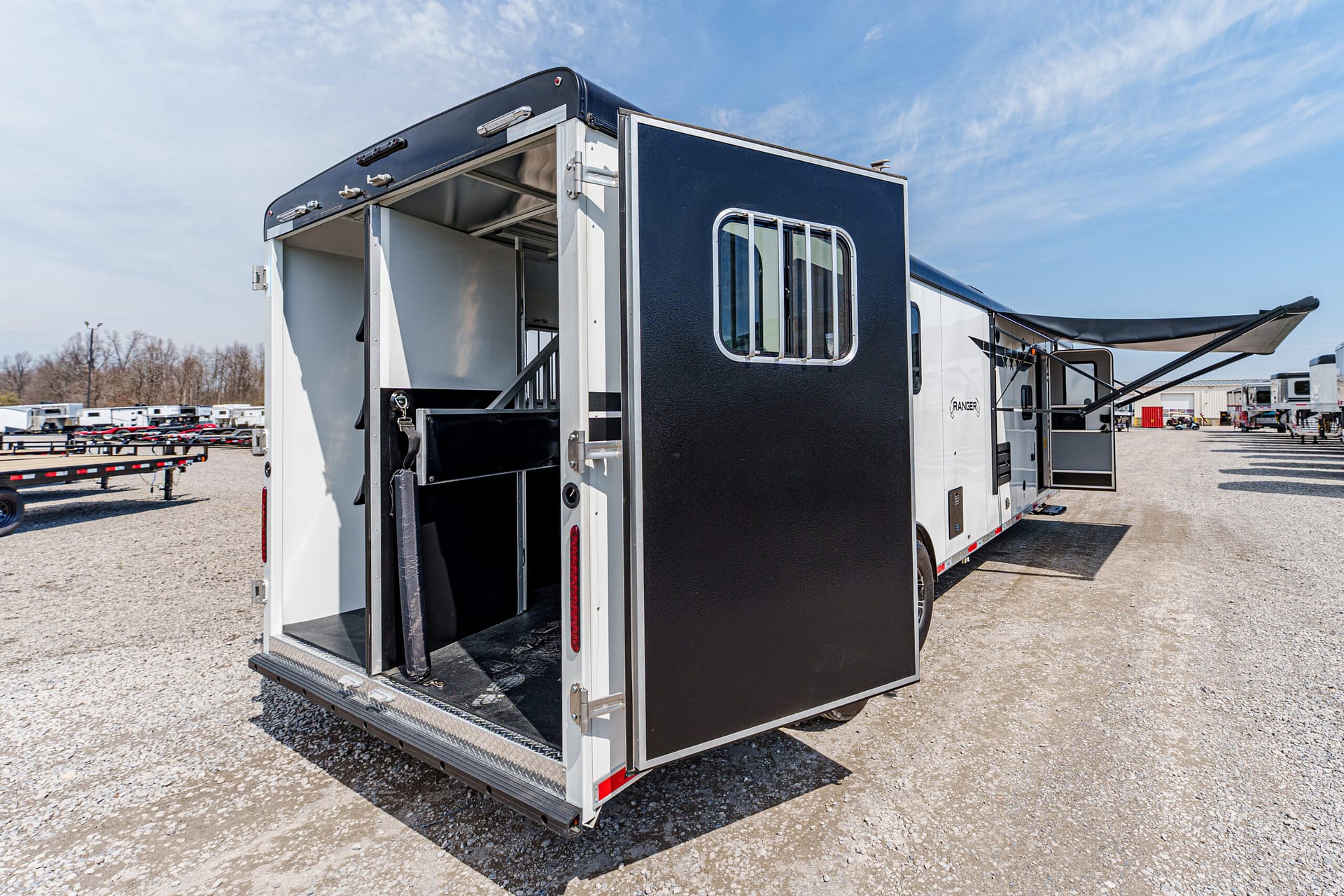 White and black horse trailer with the rear ramp down, revealing the tack area and partition inside, parked on a gravel lot under a blue sky.