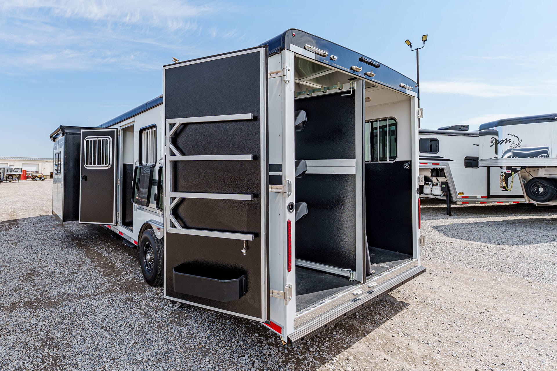 Rear of a black-and-silver horse trailer with open doors, revealing interior stalls and divider rails on a gravel lot.