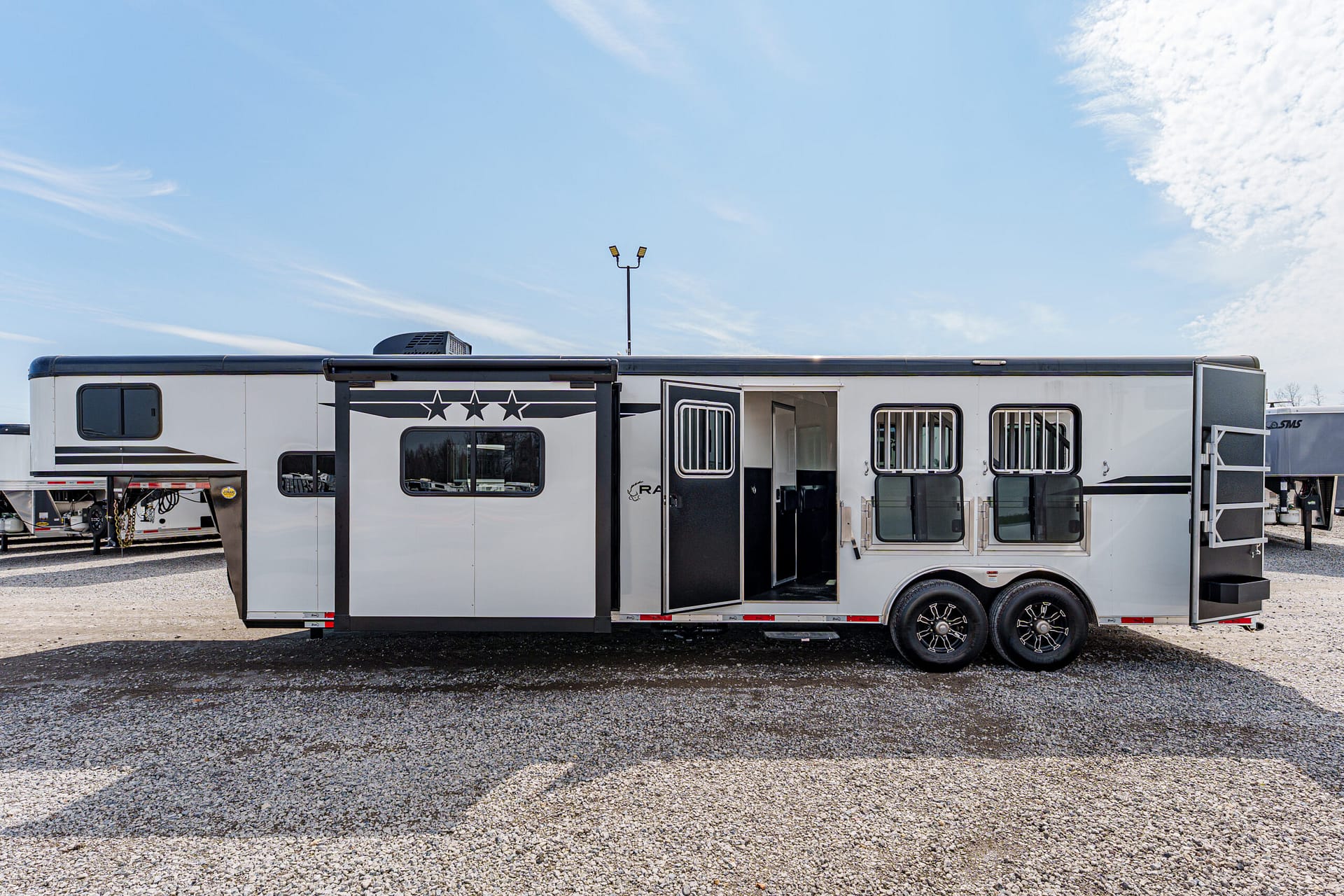 White horse trailer with black trim and star decals, open side door, parked on gravel under a blue sky